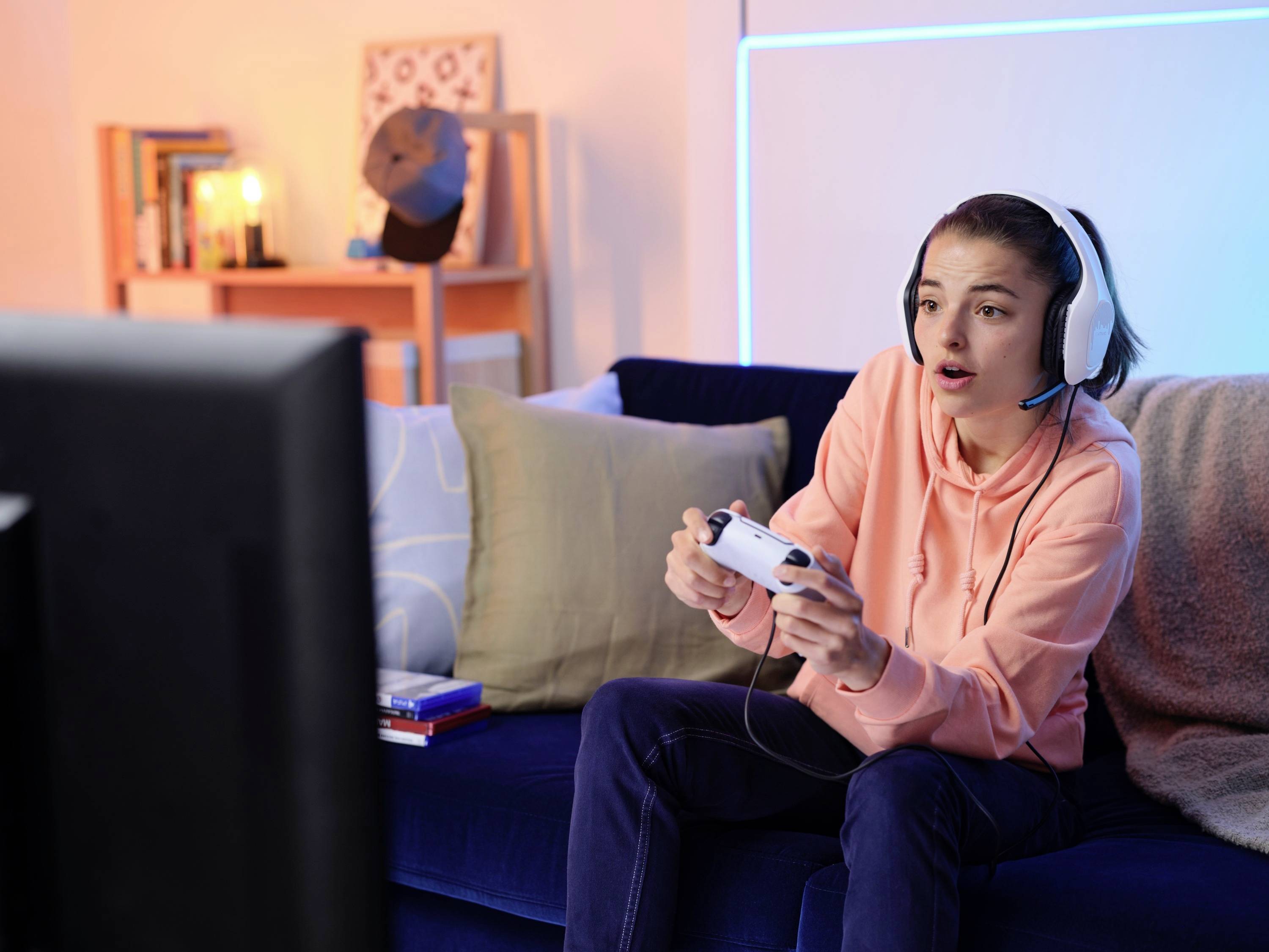 Eine junge Person sitzt auf einem Sofa, spielt Videospiele und trägt ein Headset. Im Hintergrund ein Regal mit Büchern und Dekor.