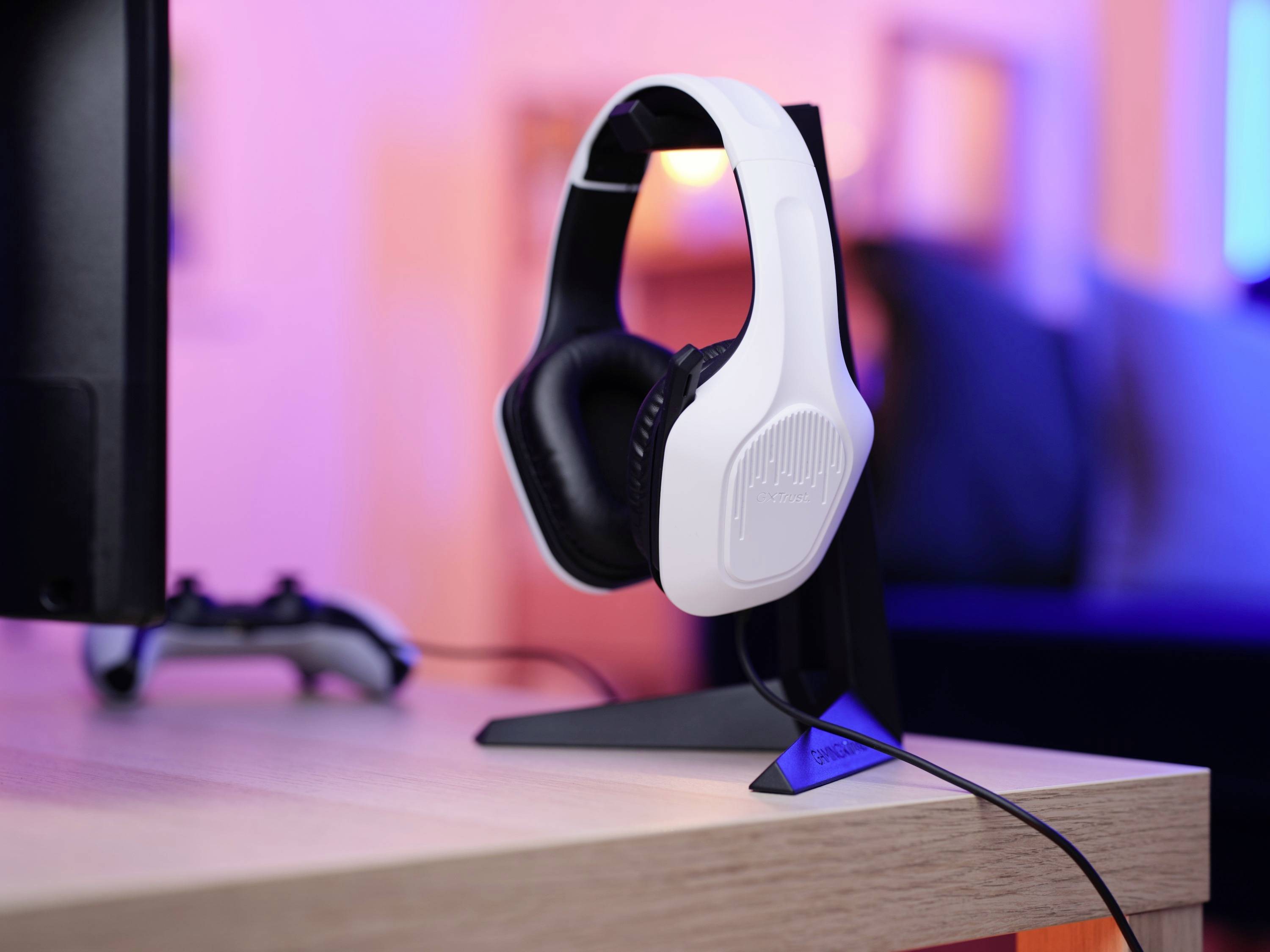 Ein weiß-schwarzer Gaming-Kopfhörer steht auf einem Ständer neben einem Bildschirm und einem Controller auf einem Tisch in beleuchteter Umgebung.