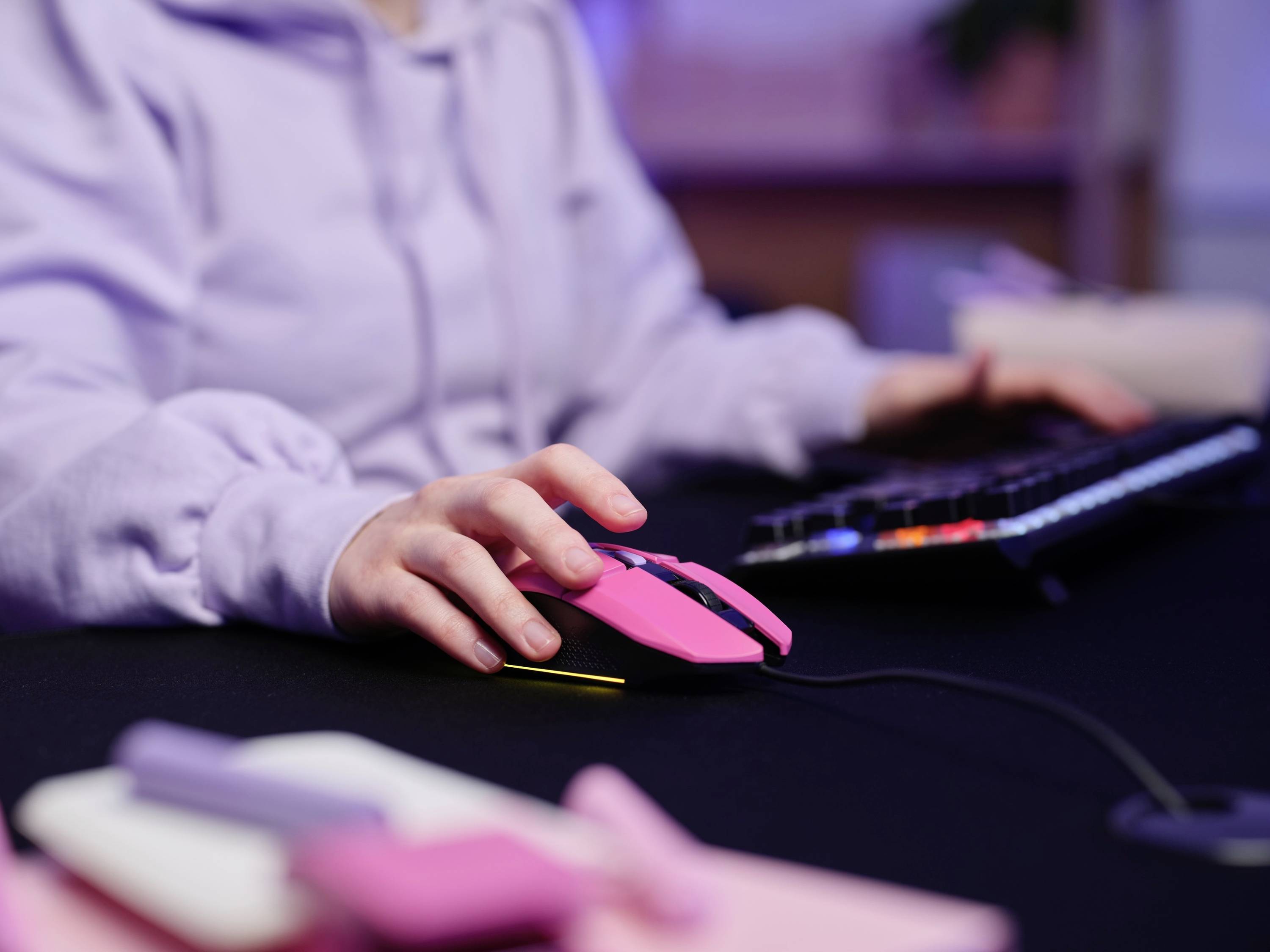 Person in grauem Hoodie verwendet eine pinke Computermaus und Tastatur auf schwarzem Tisch. Vorne liegen unscharf Schreibwaren.