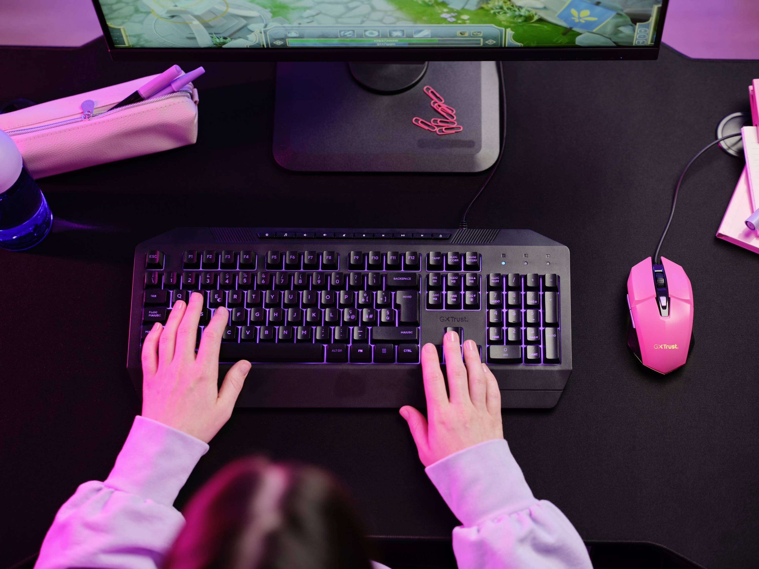 Eine Person tippt auf einer beleuchteten Tastatur vor einem Monitor. Eine rosa Maus und Schreibutensilien liegen auf dem Tisch.