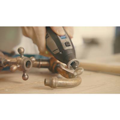 Hand hält ein Drehwerkzeug, das ein Metallrohr auf einem Holztisch bearbeitet. Im Hintergrund unscharf, Fokus auf das Werkzeug und Rohr.