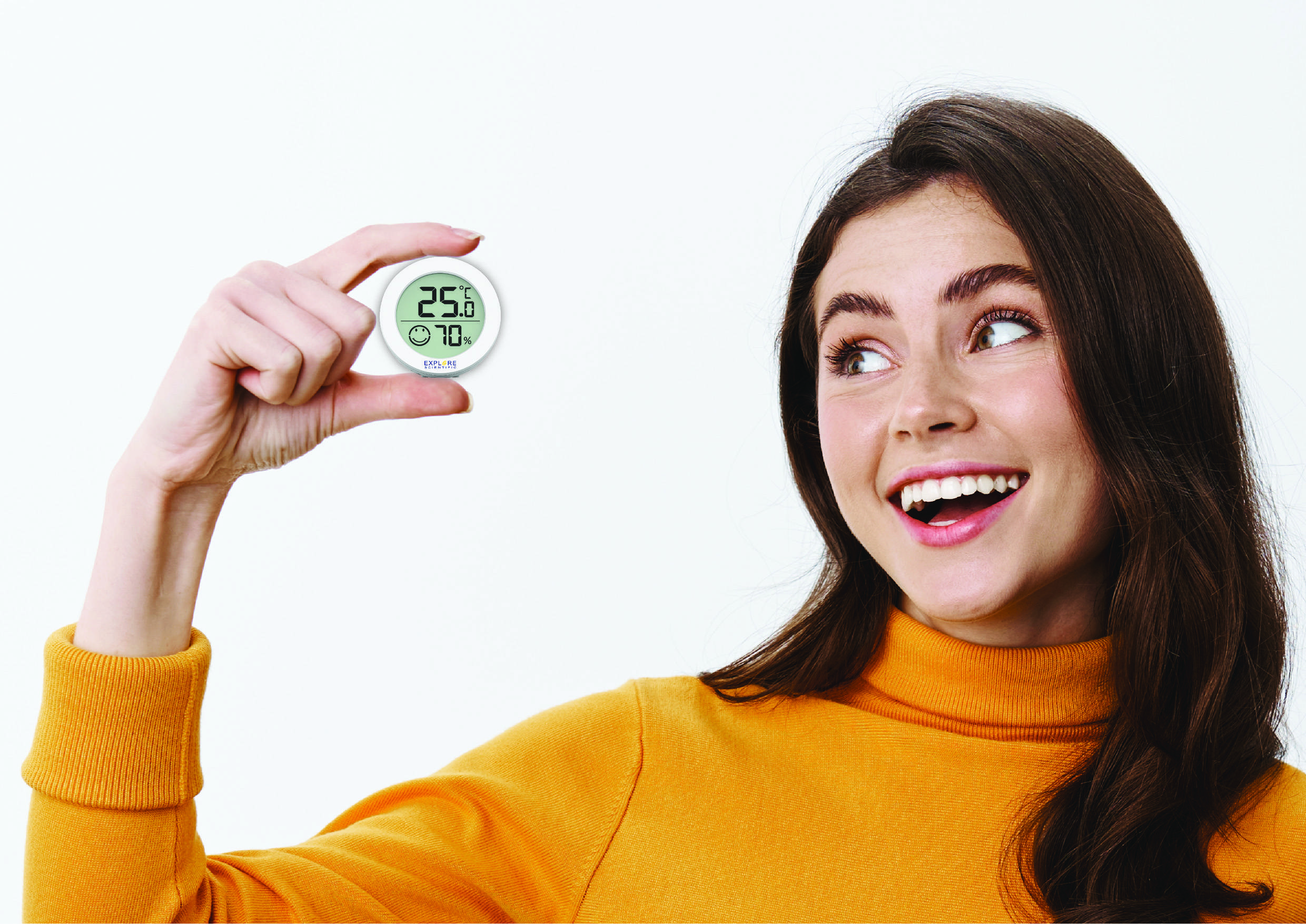 Frau in orangem Pulli hält ein rundes digitales Thermometer und Hygrometer, zeigt 25,6°C und 70% Luftfeuchtigkeit, lächelnd.