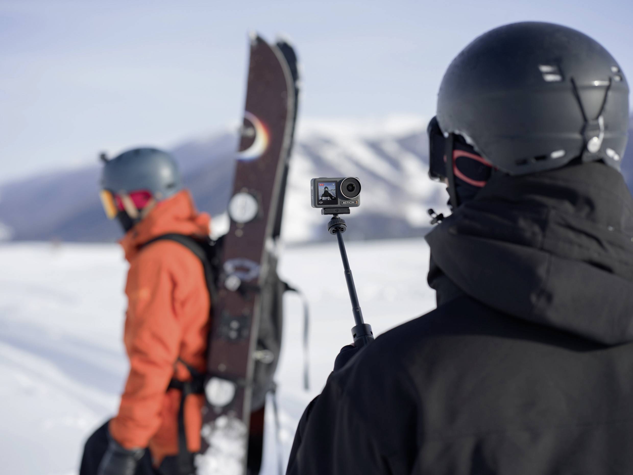 Zwei Personen in Winterausrüstung auf verschneitem Berg. Eine trägt ein Snowboard, die andere filmt mit einer Action-Kamera.