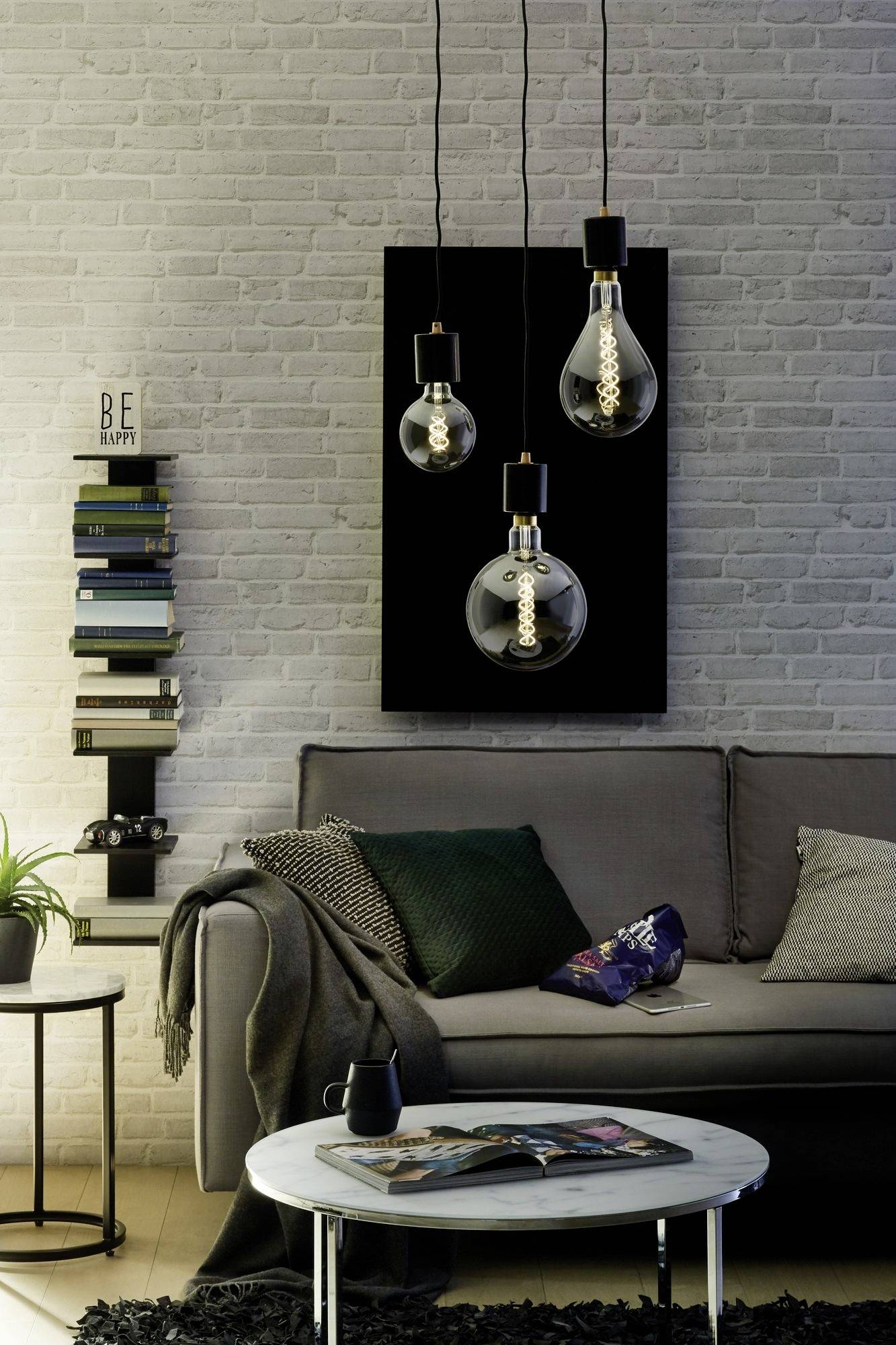 Wohnzimmer mit grauem Sofa, Kissen, Bücherstapel, Pflanze und rundem Tisch. Drei große Glühbirnen hängen als dekorative Beleuchtung.