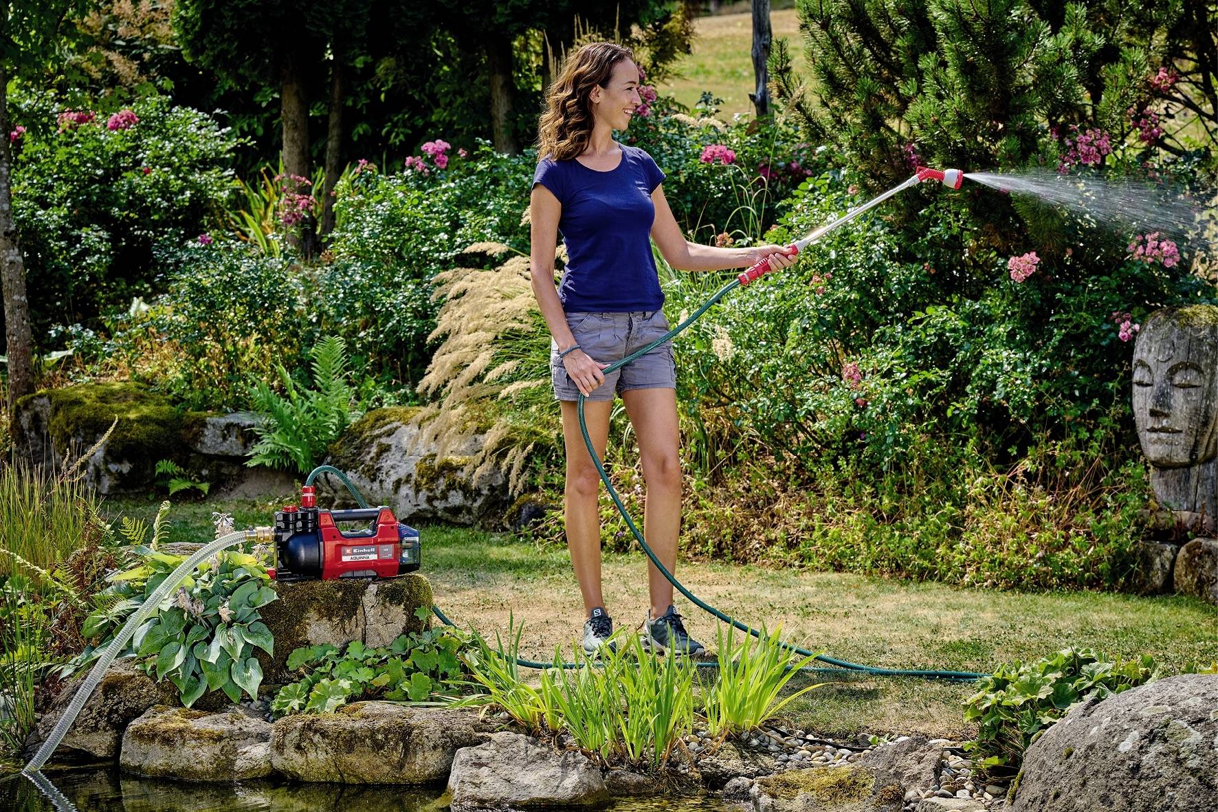 Eine Frau wässert mit einem Gartenschlauch Pflanzen in einem blühenden Garten mit Teich und Buddha-Statue im Hintergrund.