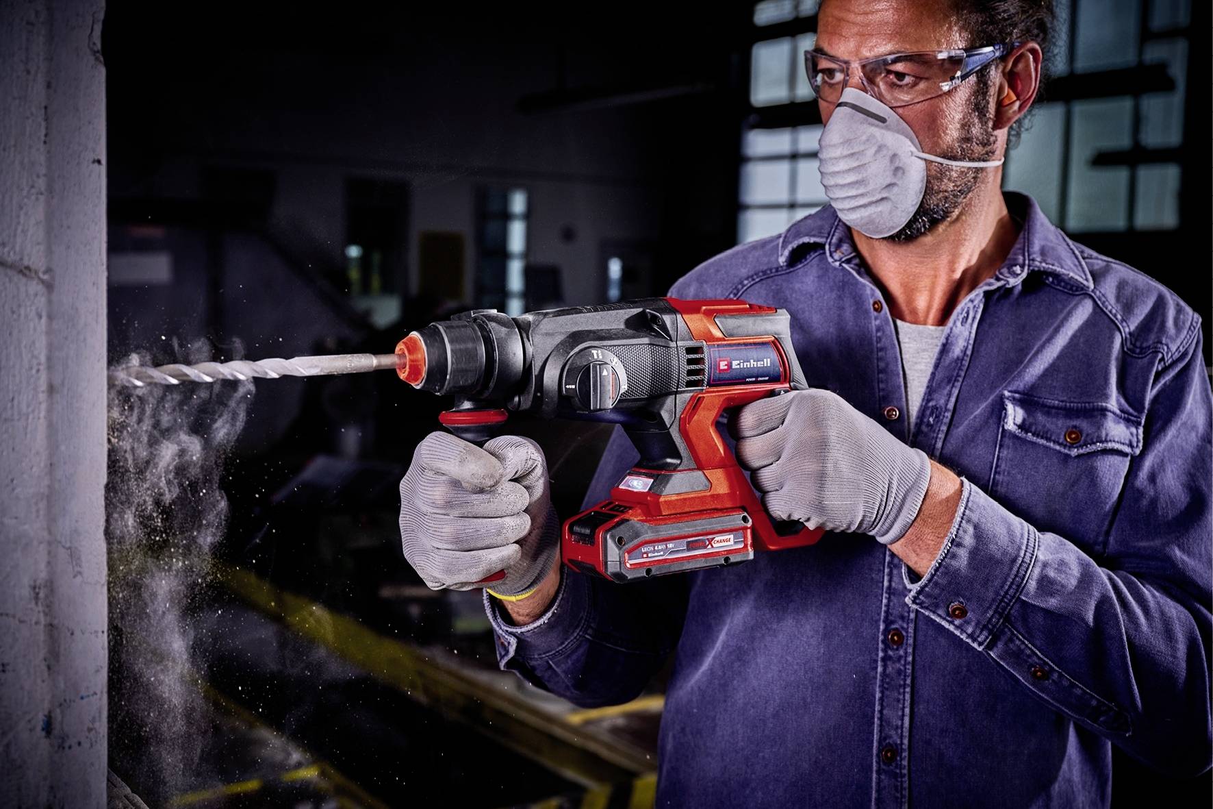 Ein Mann trägt Schutzbrille und Maske, während er mit einem elektrischen Bohrhammer in eine Betonwand bohrt.