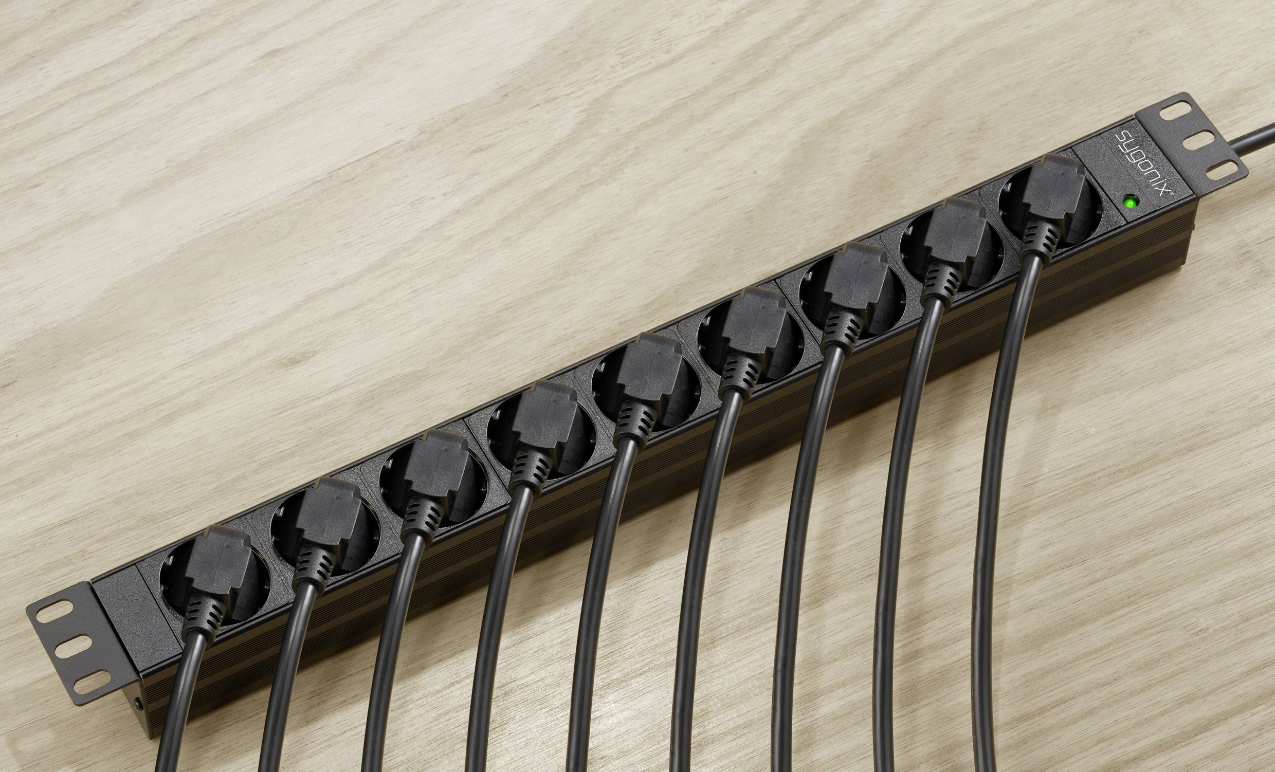 Eine schwarze Steckdosenleiste mit zehn eingesteckten schwarzen Kabeln liegt auf einem Holztisch. Eine grüne LED leuchtet.