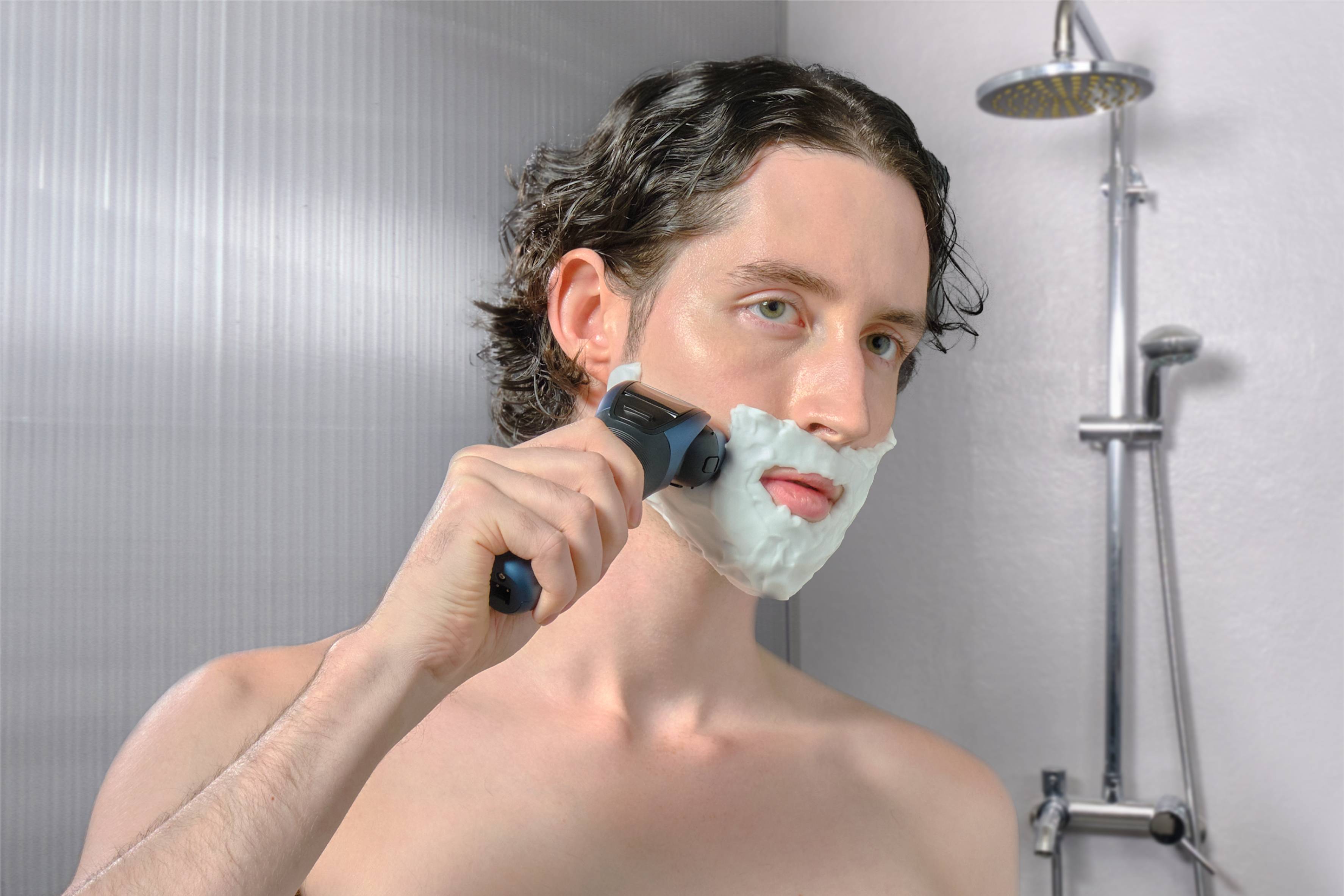 Ein Mann rasiert sich mit einem Elektrorasierer im Badezimmer. Er hat Schaum auf dem Gesicht und ist vor einer Dusche stehend.