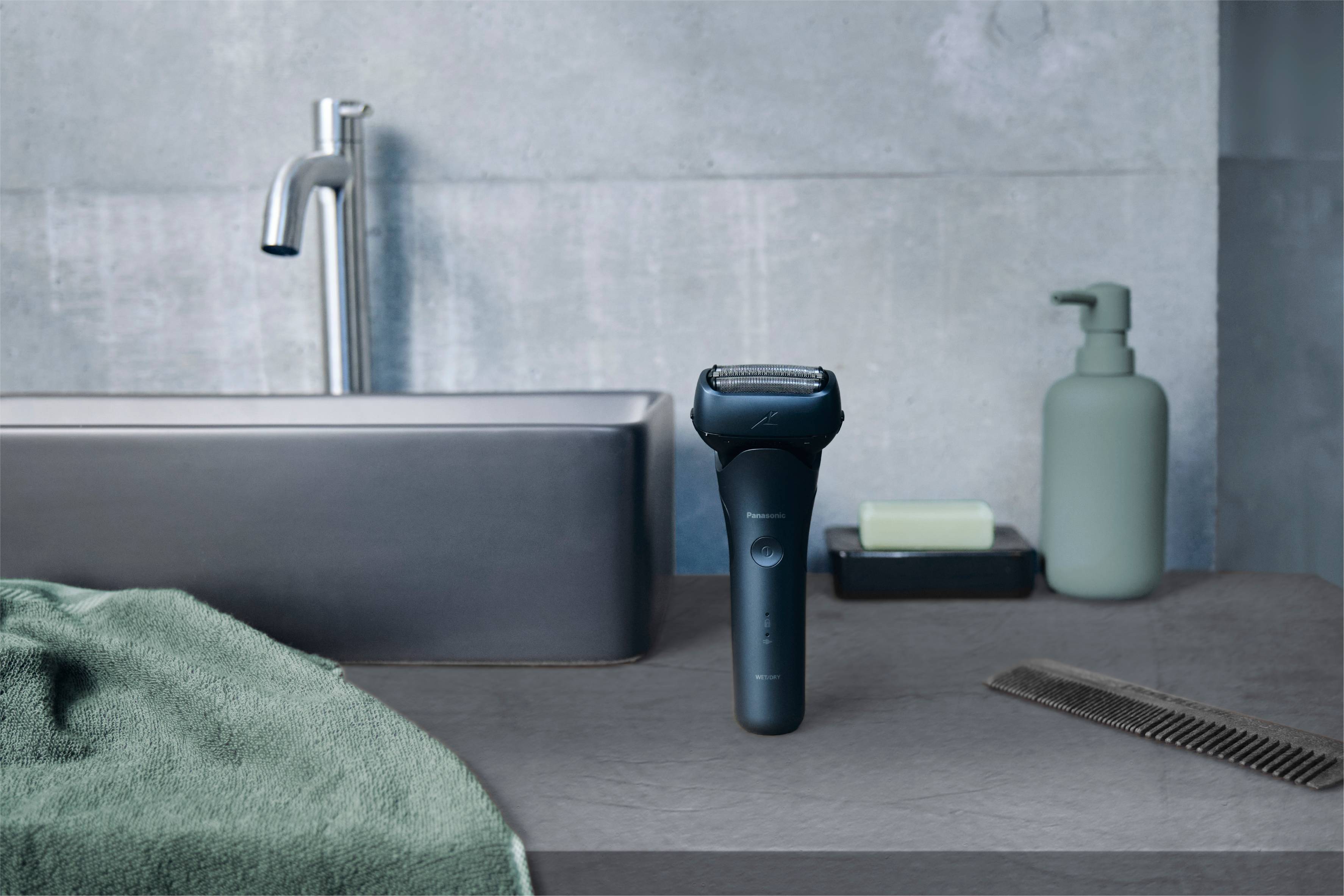 Elektrorasierer auf grauer Badezimmer-Theke neben Waschbecken, Seifenspender und Kamm.