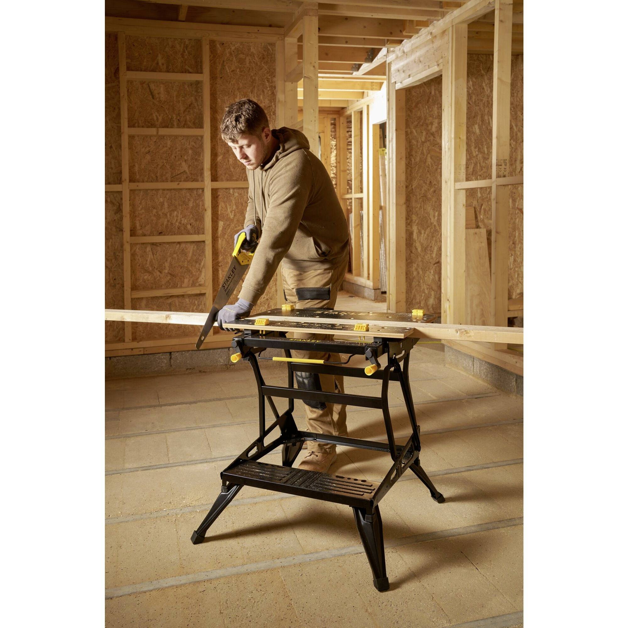 Ein Mann in einem im Bau befindlichen Raum, sägt mit einer Säge in einem Winkel auf einem Klapptisch Holz. Hintergrund: Holzwände.