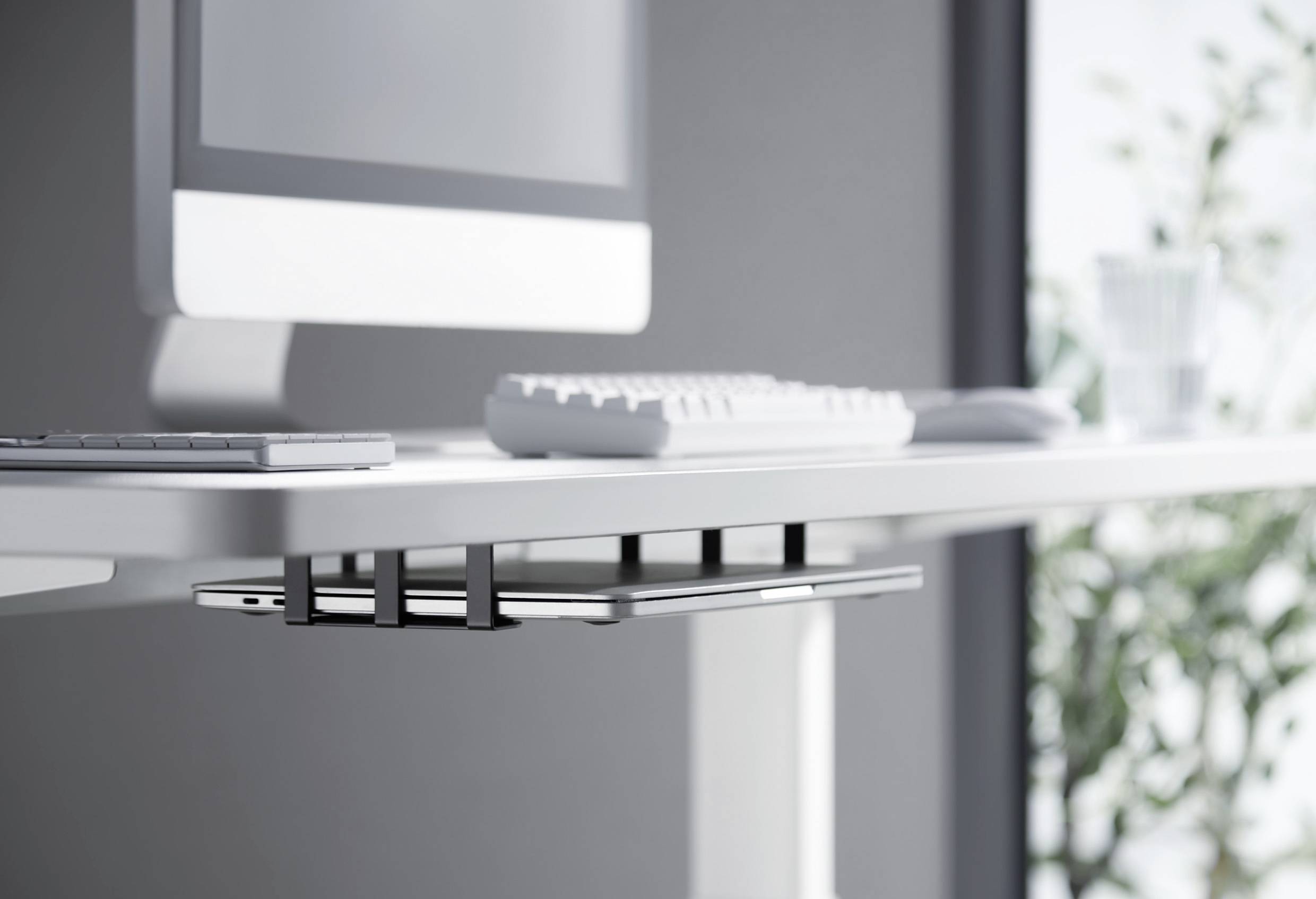 Ein moderner Schreibtisch mit einem Computerbildschirm, einer Tastatur und einem Tablet in einer Halterung darunter. Im Hintergrund verschwommene Pflanzen.