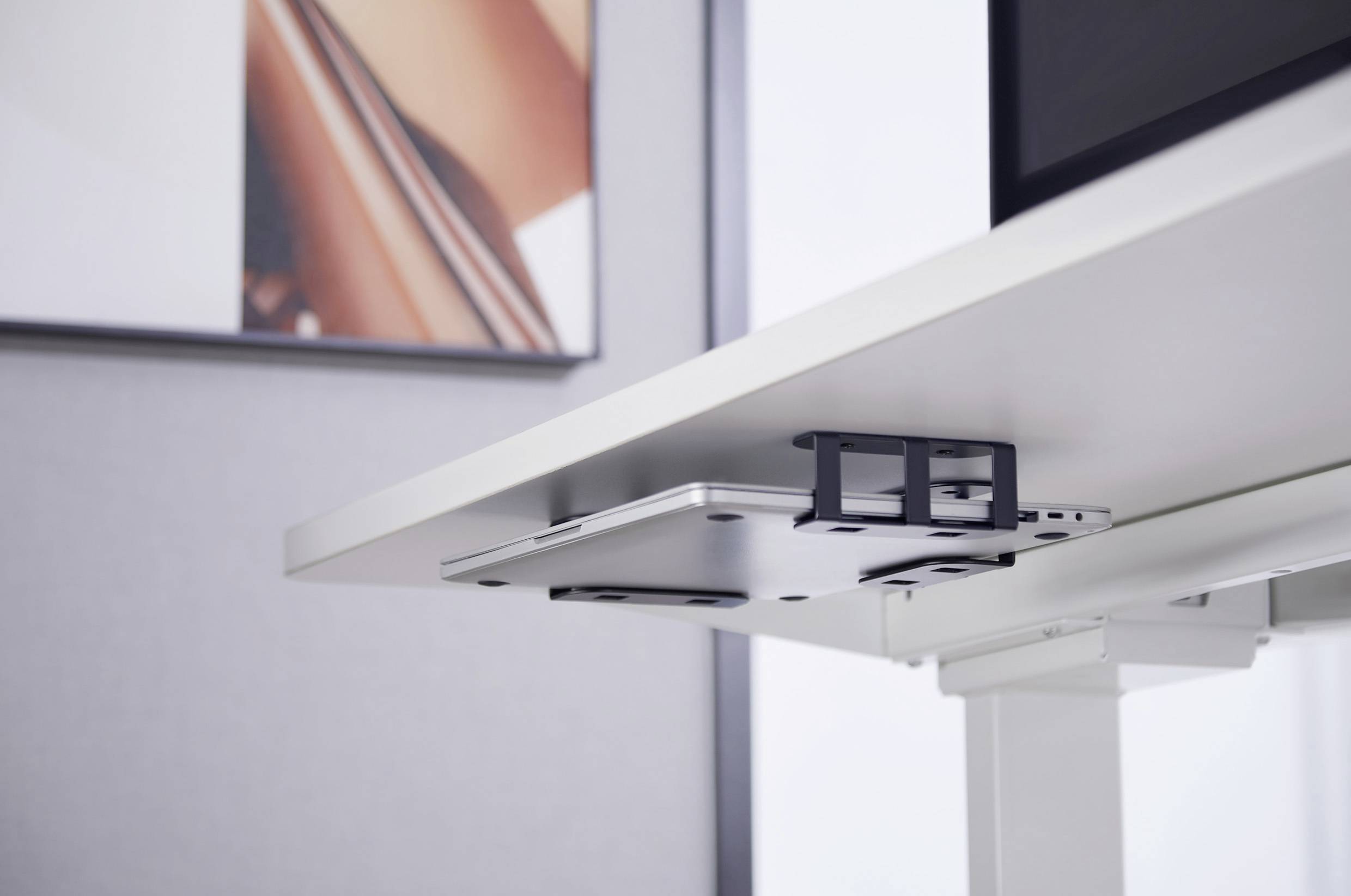 Ein Laptop ist unter einem weißen Schreibtisch in einer Halterung montiert. Im Hintergrund hängt ein abstraktes Bild an der Wand.