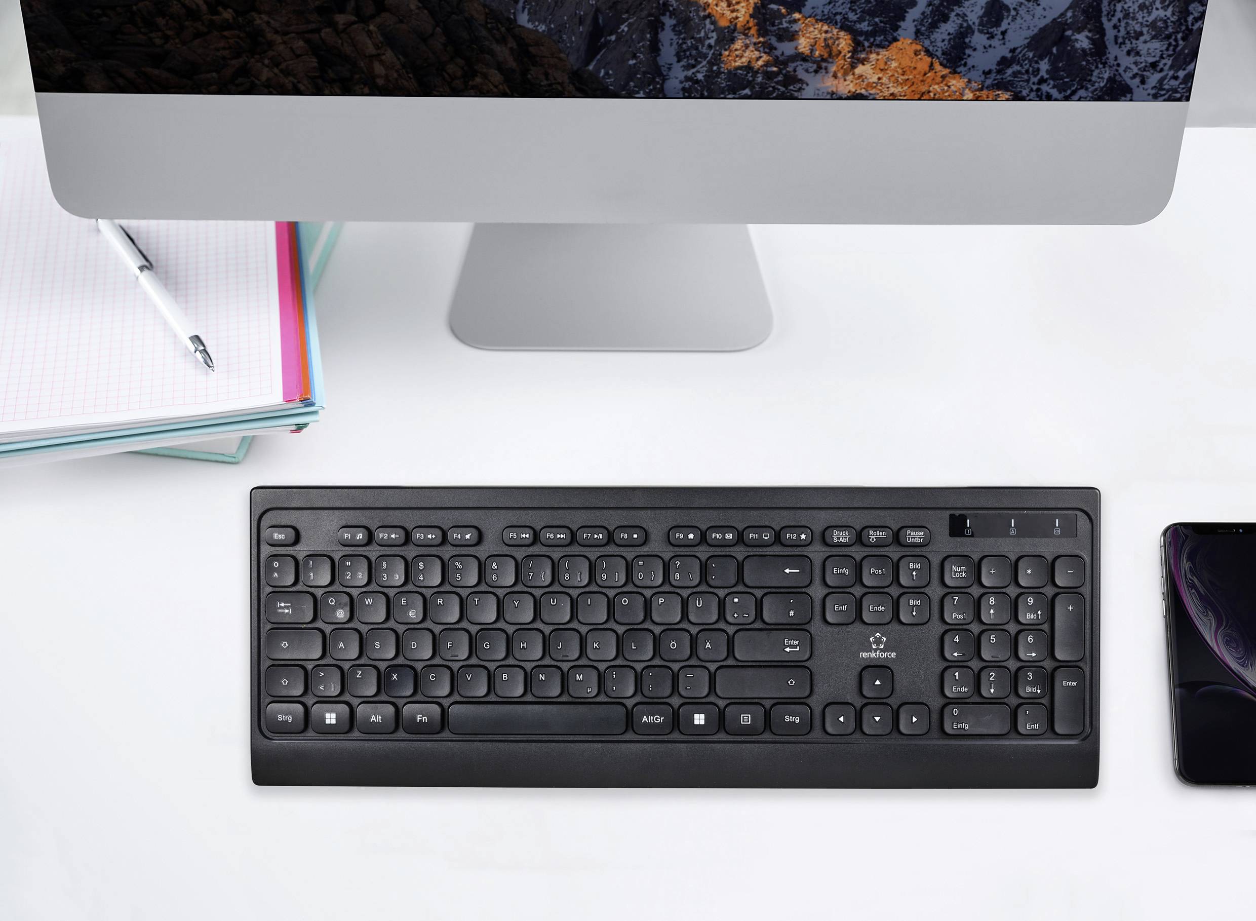 Ein schwarzes Tastatur liegt vor einem Computer auf einem Schreibtisch. Daneben befinden sich ein Notizbuch und ein Smartphone.