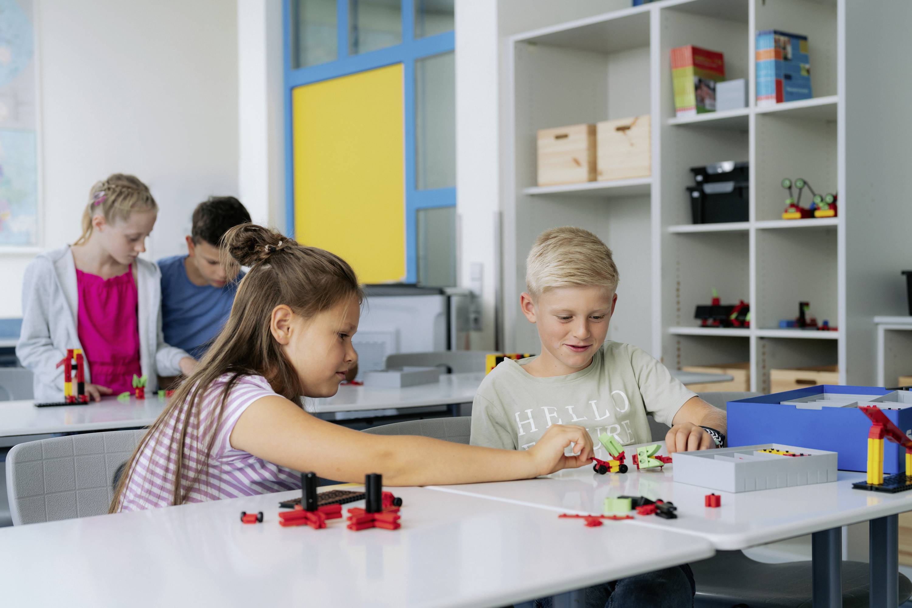 Schulkinder bauen im Klassenzimmer mit bunten Bauklötzen an einem Tisch. Im Hintergrund weitere Schüler mit ähnlichen Aktivitäten.