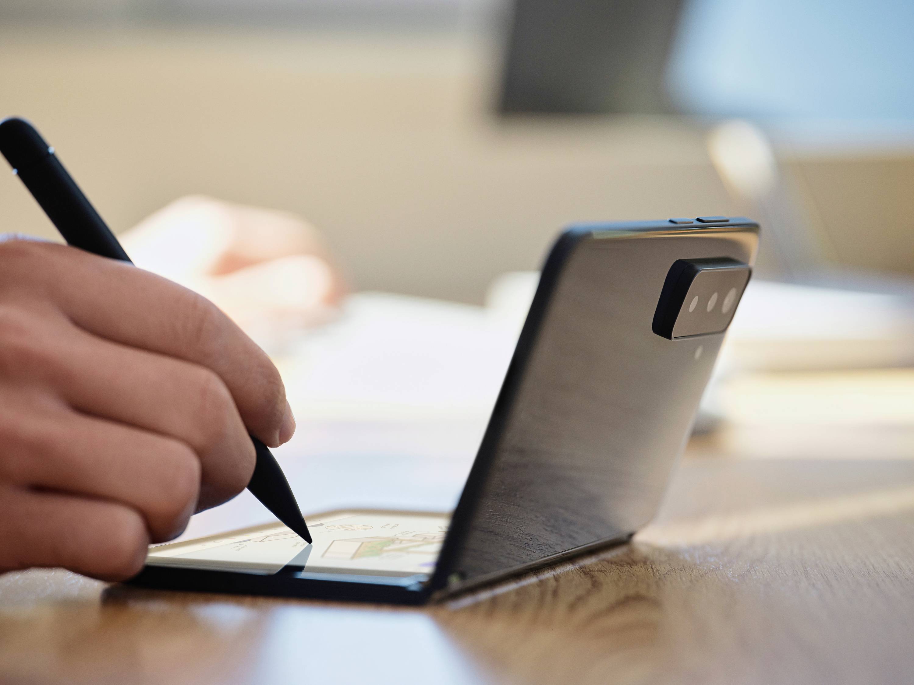 Person verwendet einen Stift auf einem klappbaren Smartphone, das auf einem Tisch steht, was auf kreative oder produktive Arbeit hinweist.