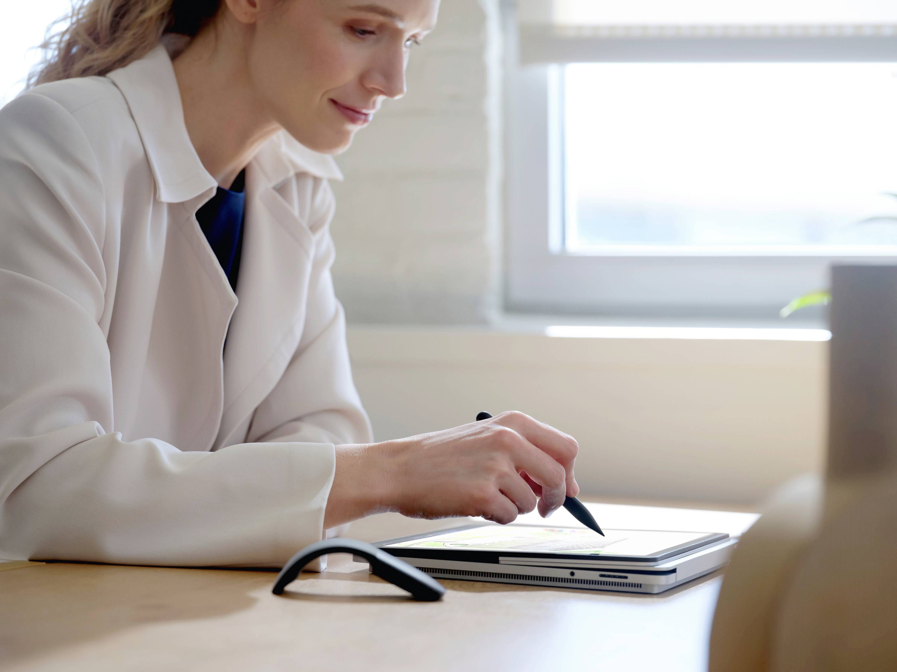 Eine Person in weißem Kittel schreibt mit einem Stift auf einem Tablet. Im Hintergrund ist ein helles Fenster zu sehen.