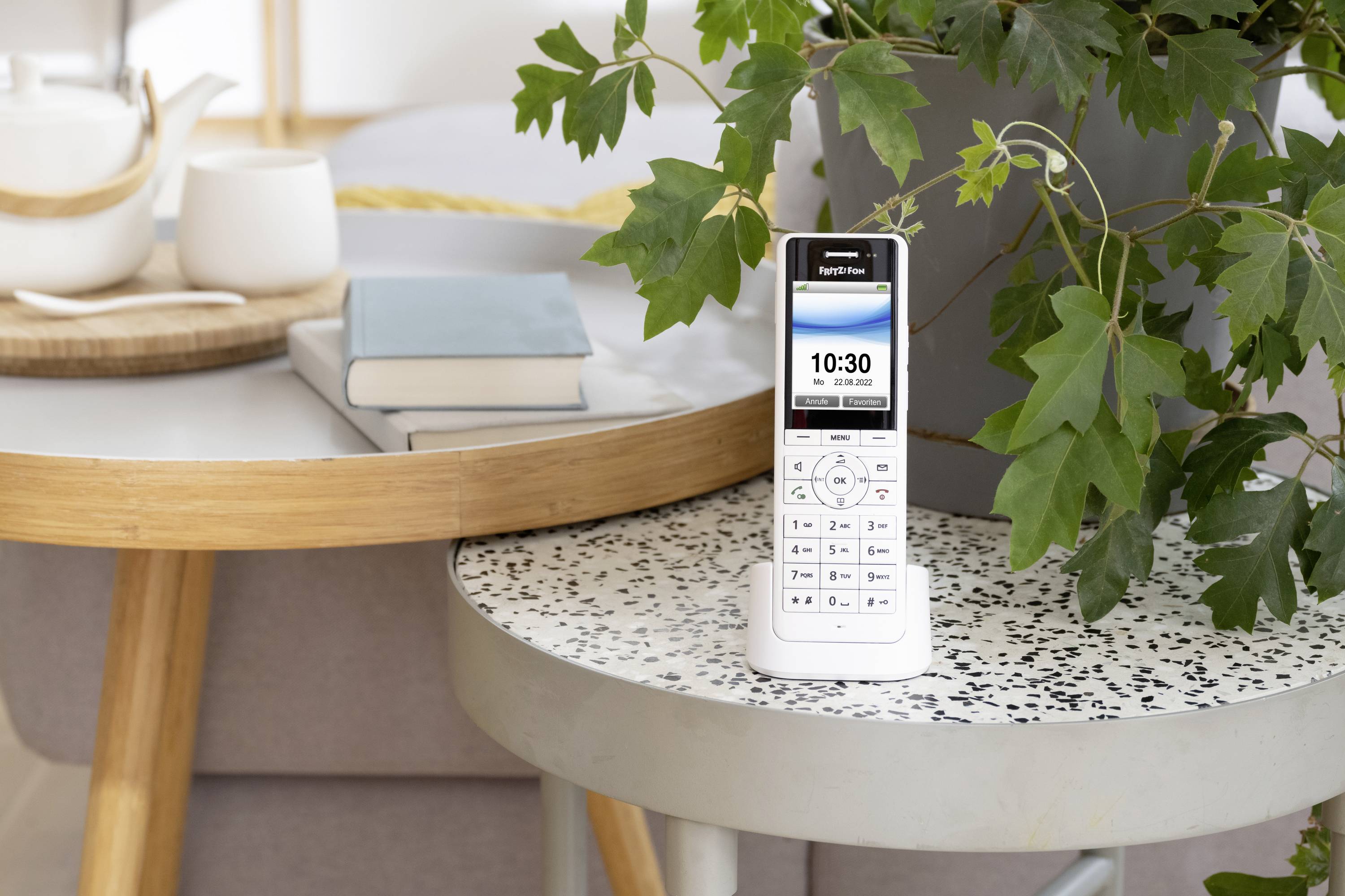 Das Bild zeigt ein IP-Telefon, dessen Mobilteil in der Basis steckt. Das Gerät steht gemeinsam mit einer Pflanze auf einem kleinen Runden Tisch. Daneben steht ein etwas größerer Tisch, auf dem sich zwei Bücher und ein Tablett mit einer Teekanne, einer Tasse und einem Löffel befinden.