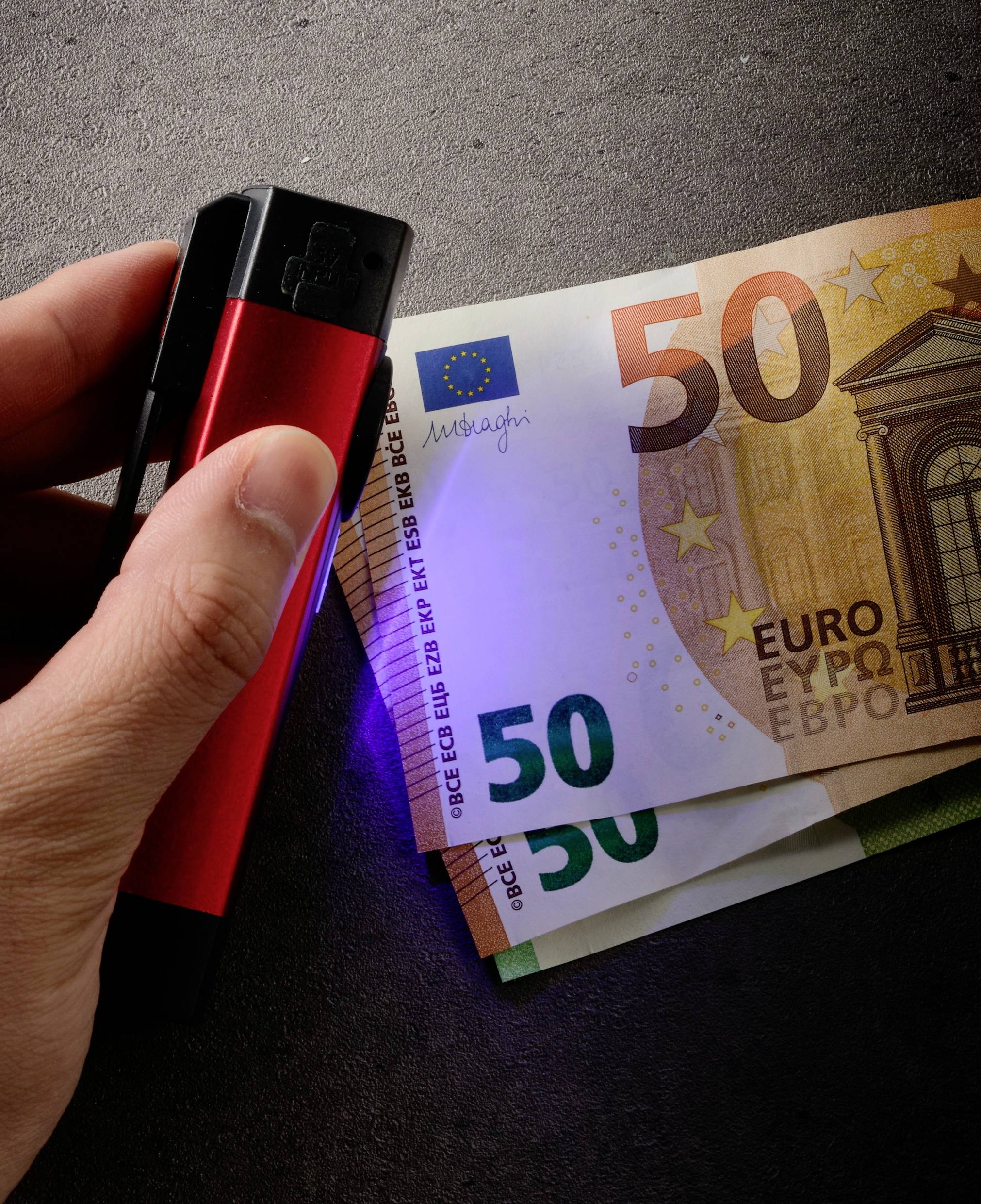 Hand hält eine UV-Lampe über einen 50-Euro-Schein, um Sicherheitsmerkmale zu überprüfen.