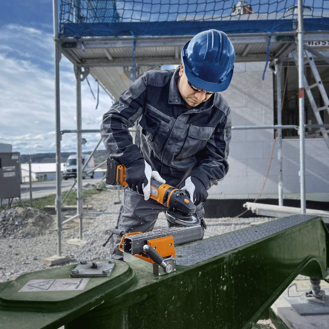 Ein Arbeiter mit Schutzkleidung und Helm benutzt eine elektrische Säge auf einer Baustelle im Freien. Im Hintergrund sind Baugerüste und ein unfertiges Gebäude zu sehen.