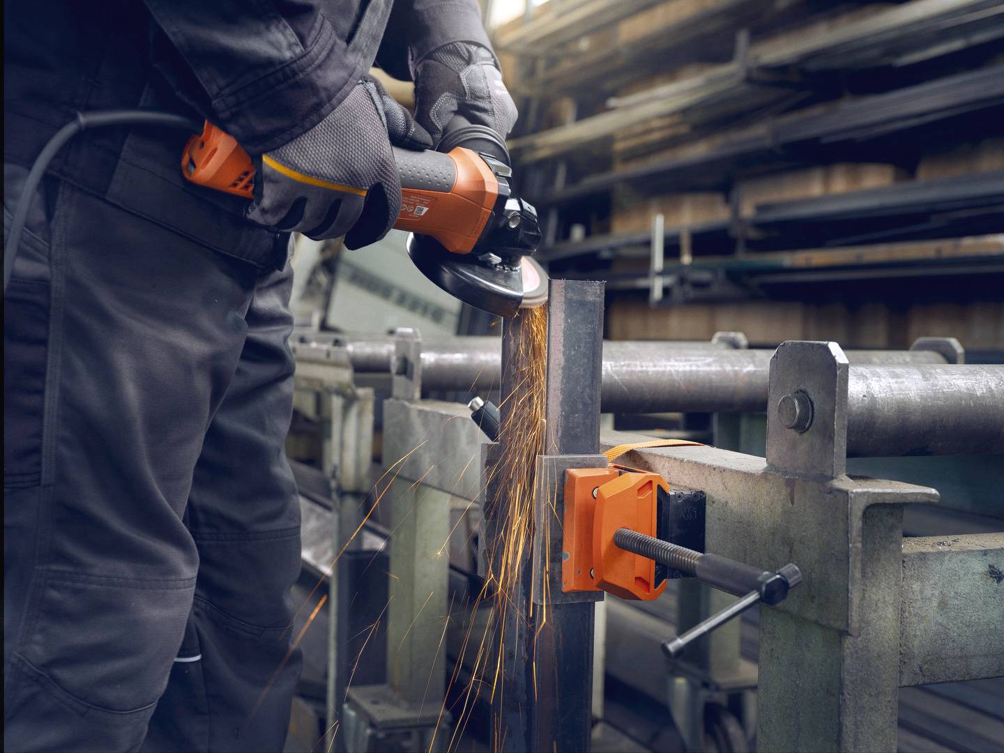 Eine Person schleift Metall mit einem Winkelschleifer, Funken fliegen. Die Umgebung ist eine Werkstatt, Metallteile sind zu sehen.