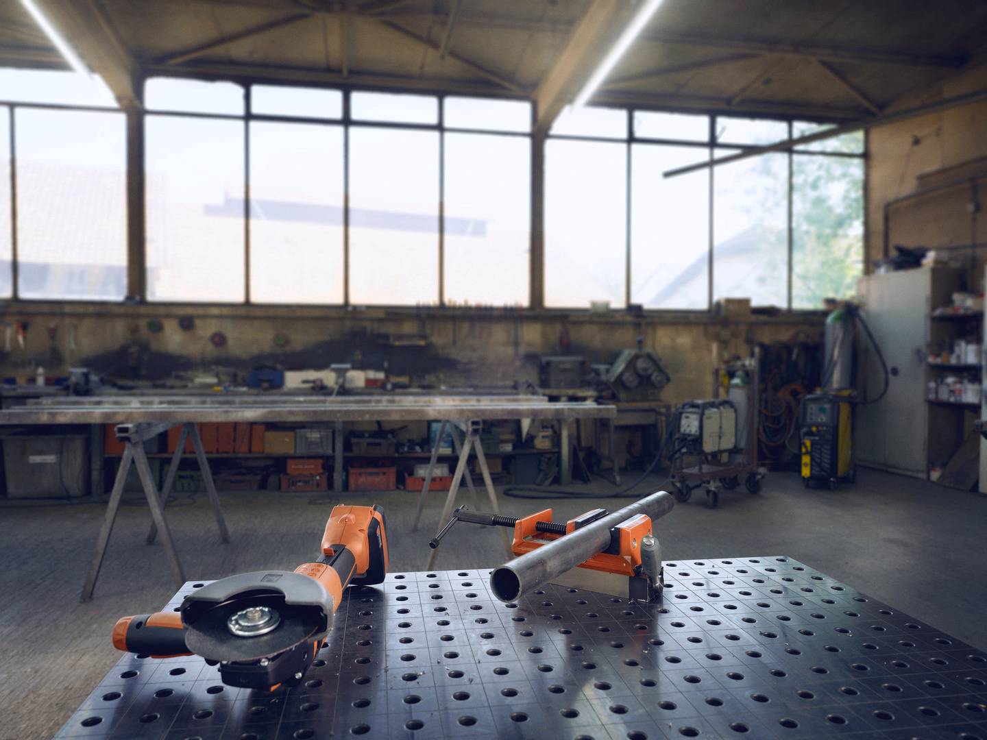 Ein Werkstatt-Arbeitsplatz mit einer Metallplatte im Vordergrund; darauf liegen ein Winkelschleifer und ein Rohr. Im Hintergrund sind Arbeitsbänke und Werkzeuge.