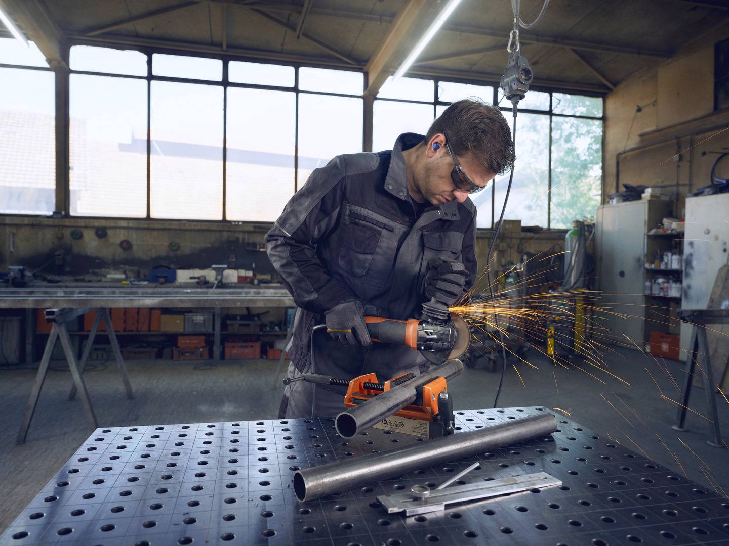 Ein Arbeiter in Schutzkleidung schleift mit einem Winkelschleifer ein Metallrohr, Funken fliegen. Hintergrund: eine Werkstatt.