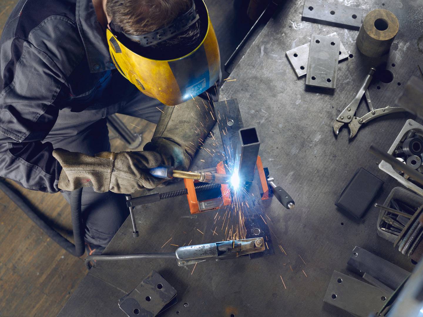 Eine Person schweißt in einer Werkstatt, trägt Schutzkleidung und Helm. Funken fliegen, während Metallteile auf der Werkbank liegen.