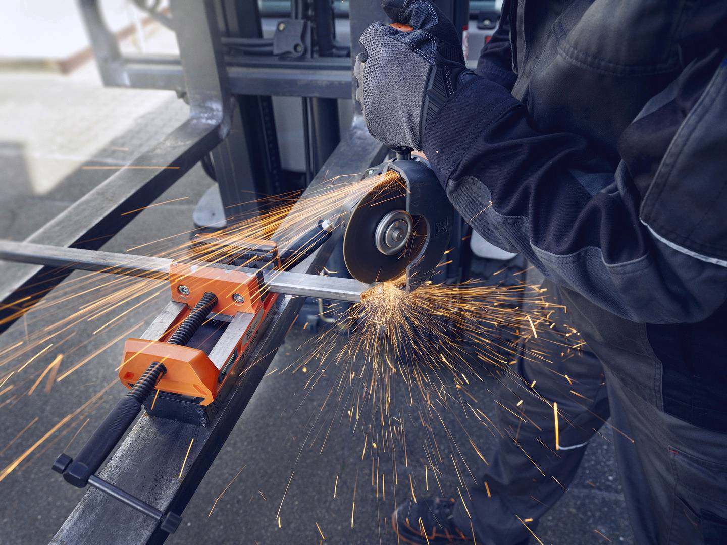 Eine Person in Arbeitskleidung schneidet Metall mit einer Schleifmaschine, es fliegen Funken. Die Szene zeigt eine Werkstattumgebung.