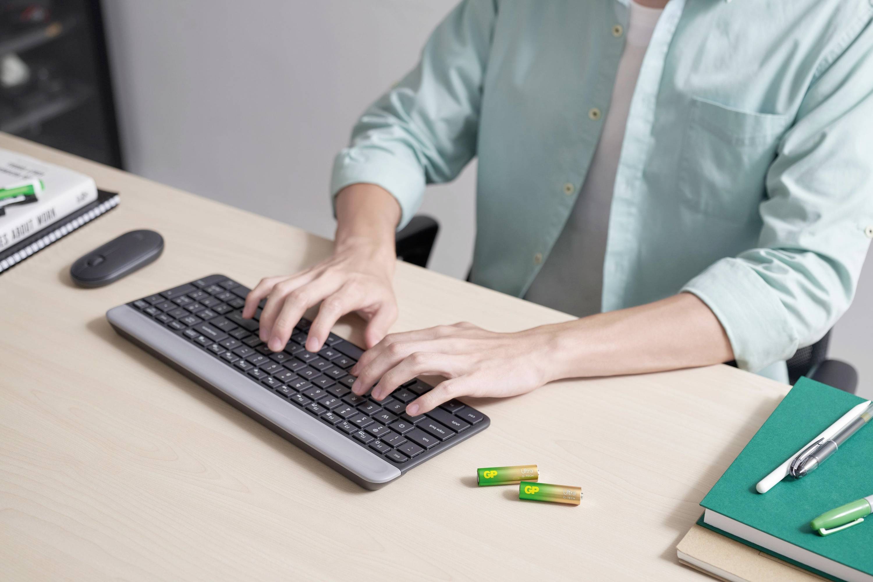Person tippt an einer Tastatur auf einem Schreibtisch; daneben liegen Stifte, Notizbuch und Batterien.