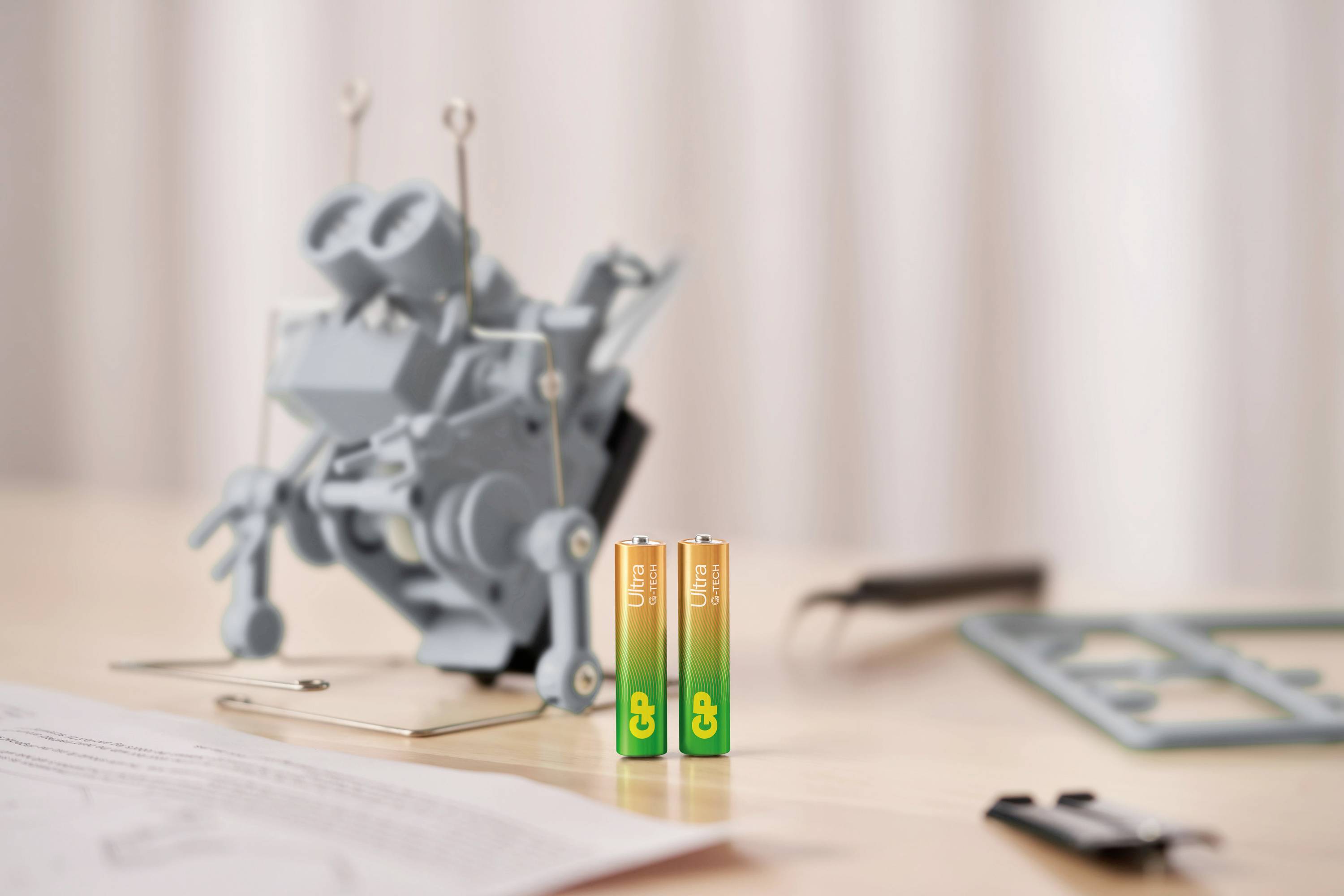 Zwei aufrechte AA-Batterien stehen vor einem unscharfen, grauen Modellbau-Roboter. Daneben liegen Papier und Werkzeuge auf dem Tisch.