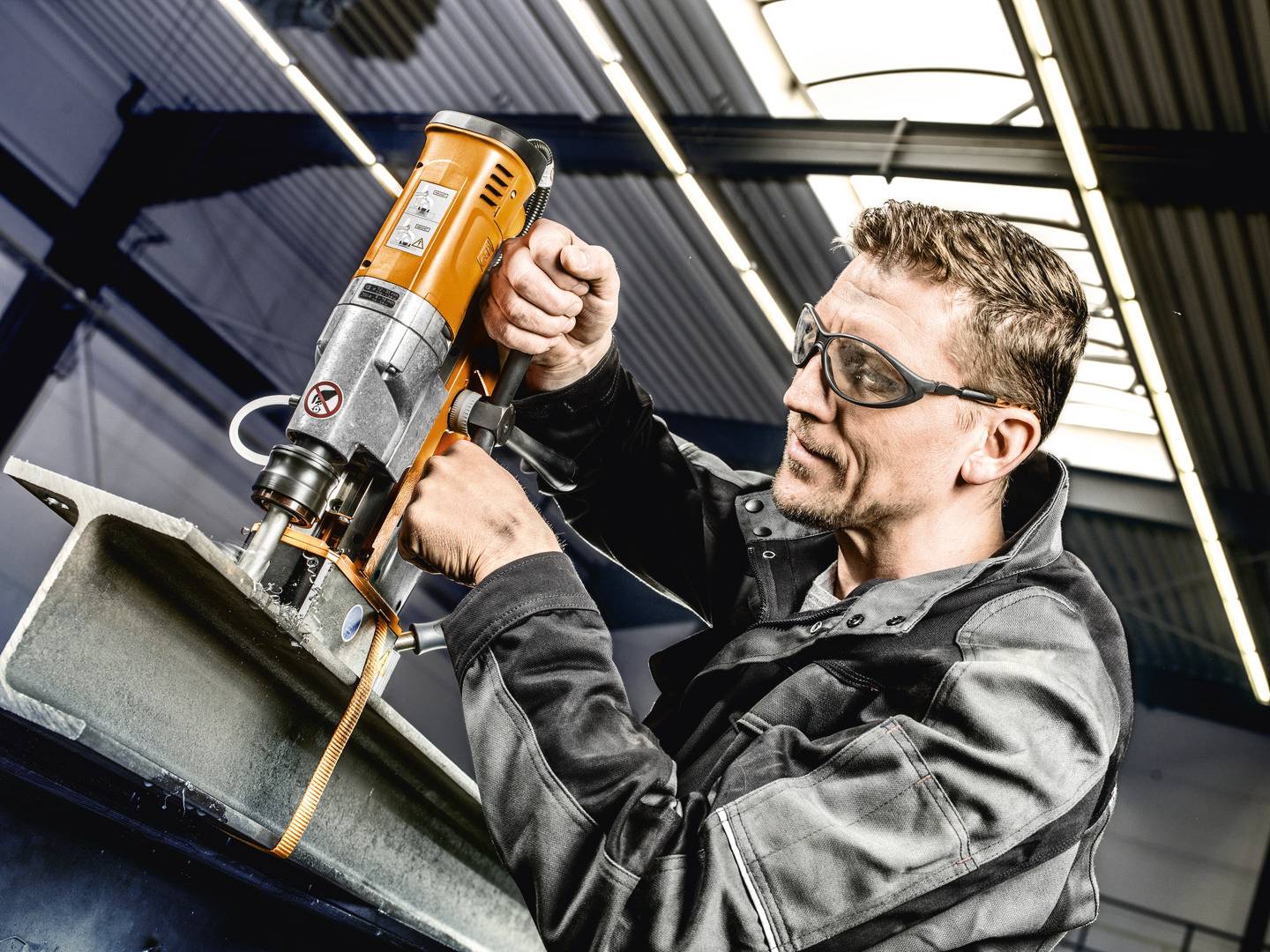 Ein Mann mit Schutzbrille bedient eine schwere Bohrmaschine in einer Werkstatt. Konzentrierter Gesichtsausdruck, industrielle Umgebung.