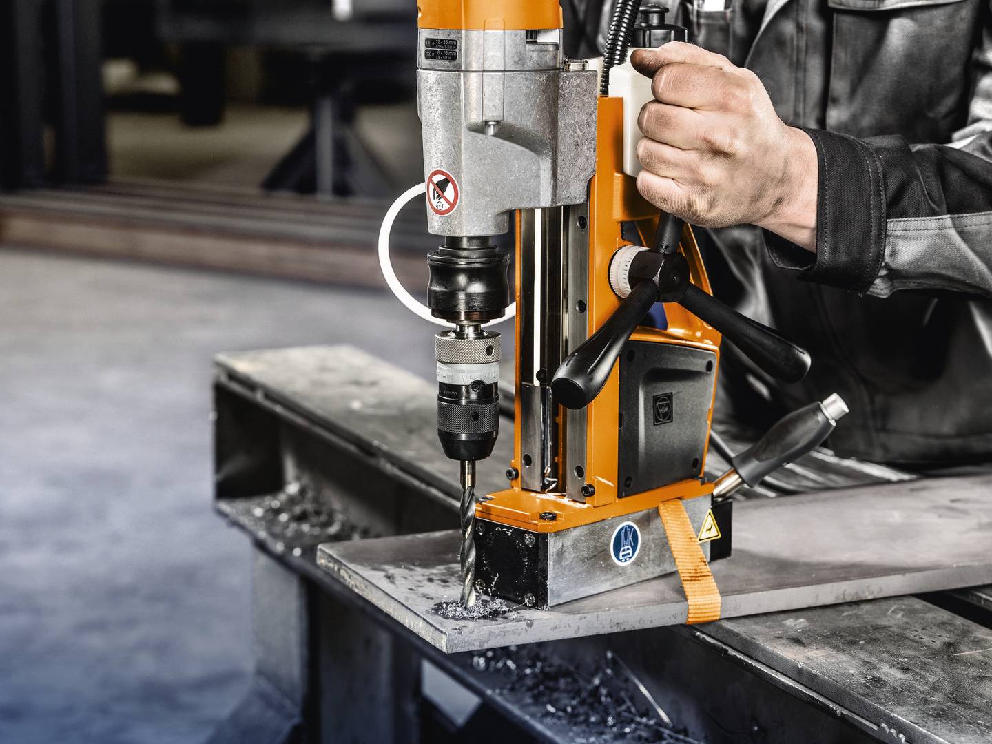 Eine Person bedient eine orangefarbene Magnetbohrmaschine auf einem Metallbalken. Metallspäne sind um die Bohrstelle sichtbar.