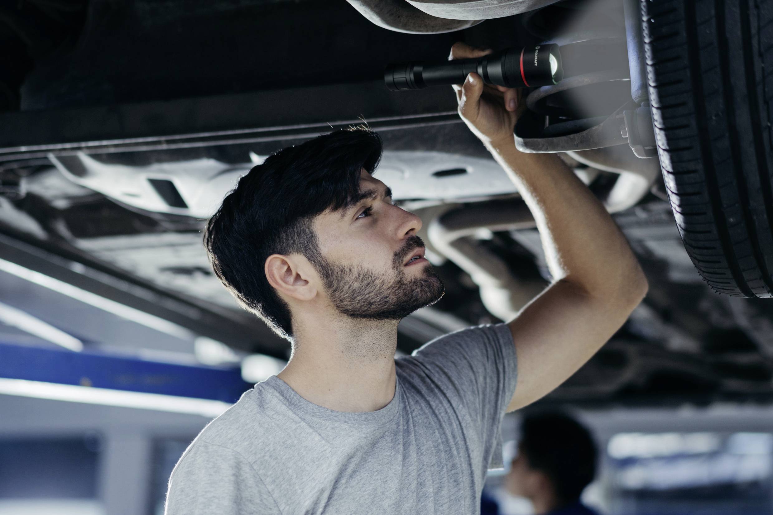 Ein Mann überprüft das Fahrwerk eines Autos in einer Werkstattumgebung. Er trägt ein graues T-Shirt und benutzt eine Lampe zur Inspektion.