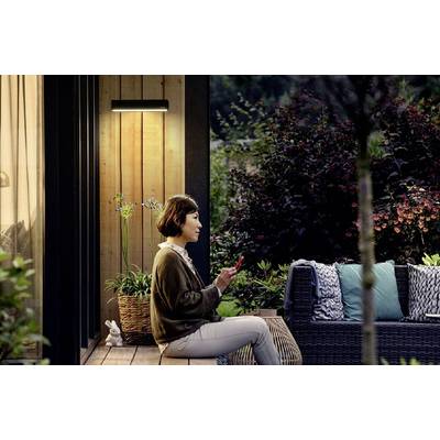 Eine Frau sitzt draußen auf einer Terrasse, umgeben von Pflanzen, und benutzt ein Smartphone. Es ist Abend und ein Wandlicht leuchtet.