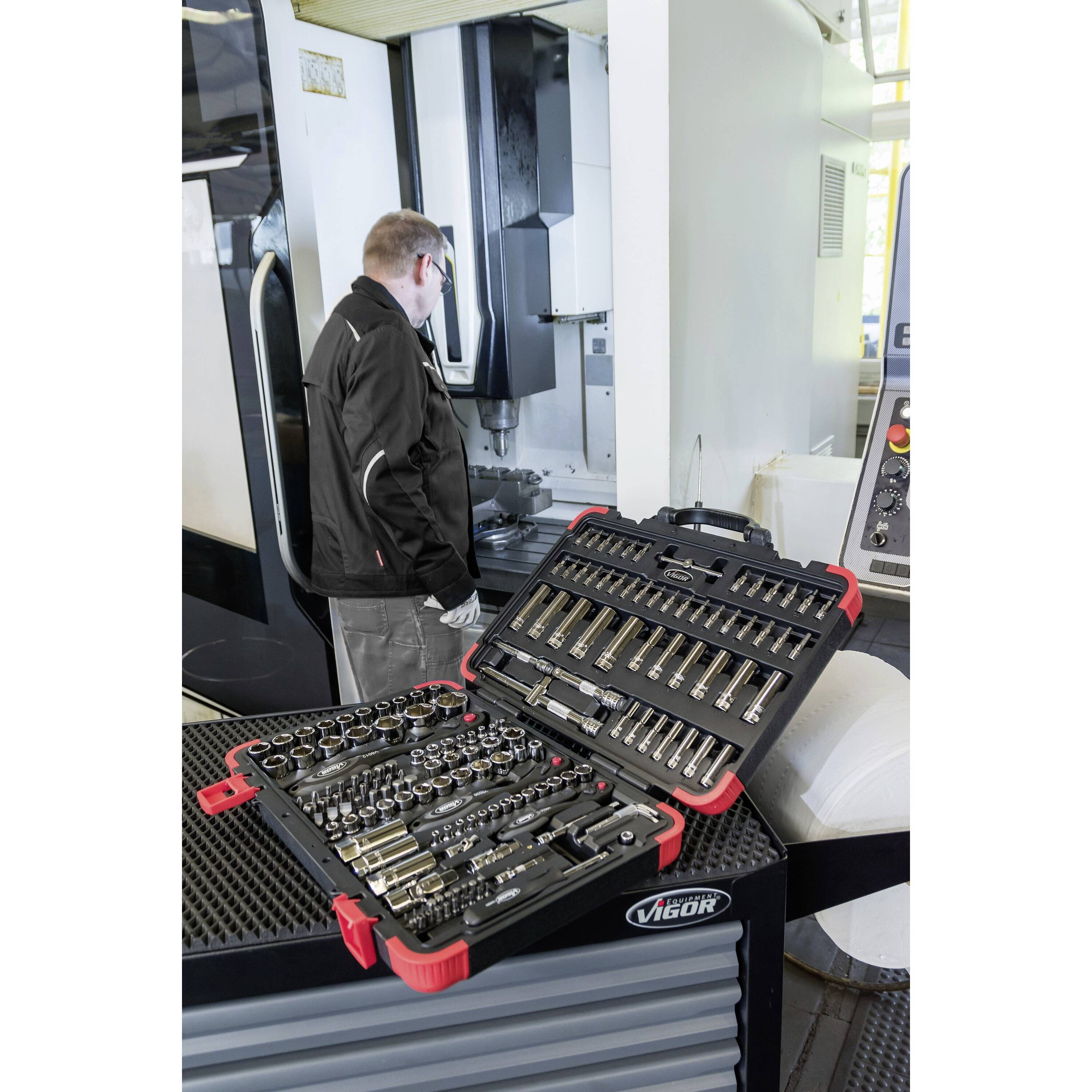 Eine Person steht in einer Werkstatt neben einer Metallbearbeitungsmaschine; im Vordergrund befindet sich eine große, offene Werkzeugkiste mit verschiedenen Bohrern und Zubehörteilen.