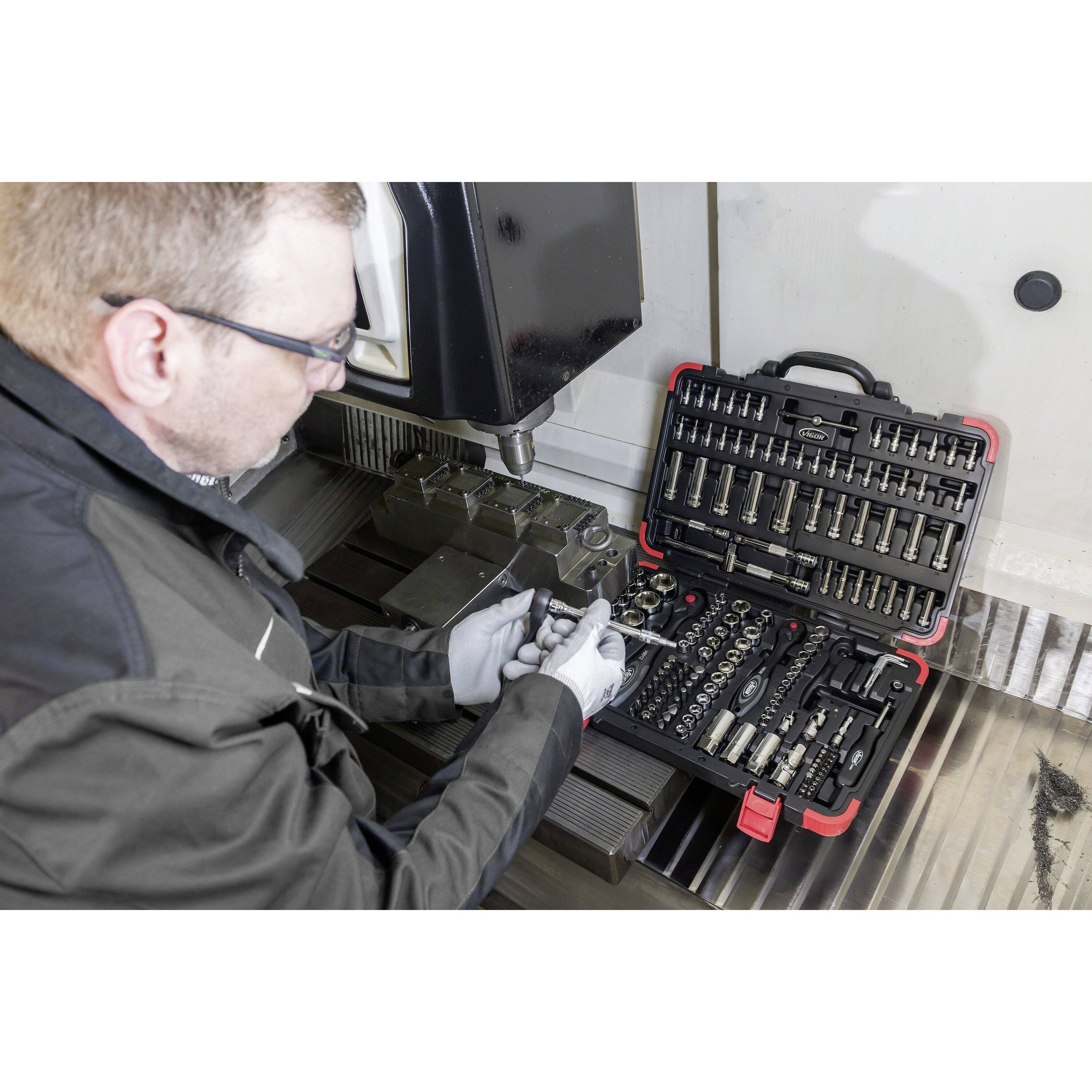 Eine Person in einer Werkstatt, die ein Werkzeug aus einer Werkzeugkiste neben einer Metallbearbeitungsmaschine auswählt und sich dabei auf Präzision und Detailgenauigkeit konzentriert.