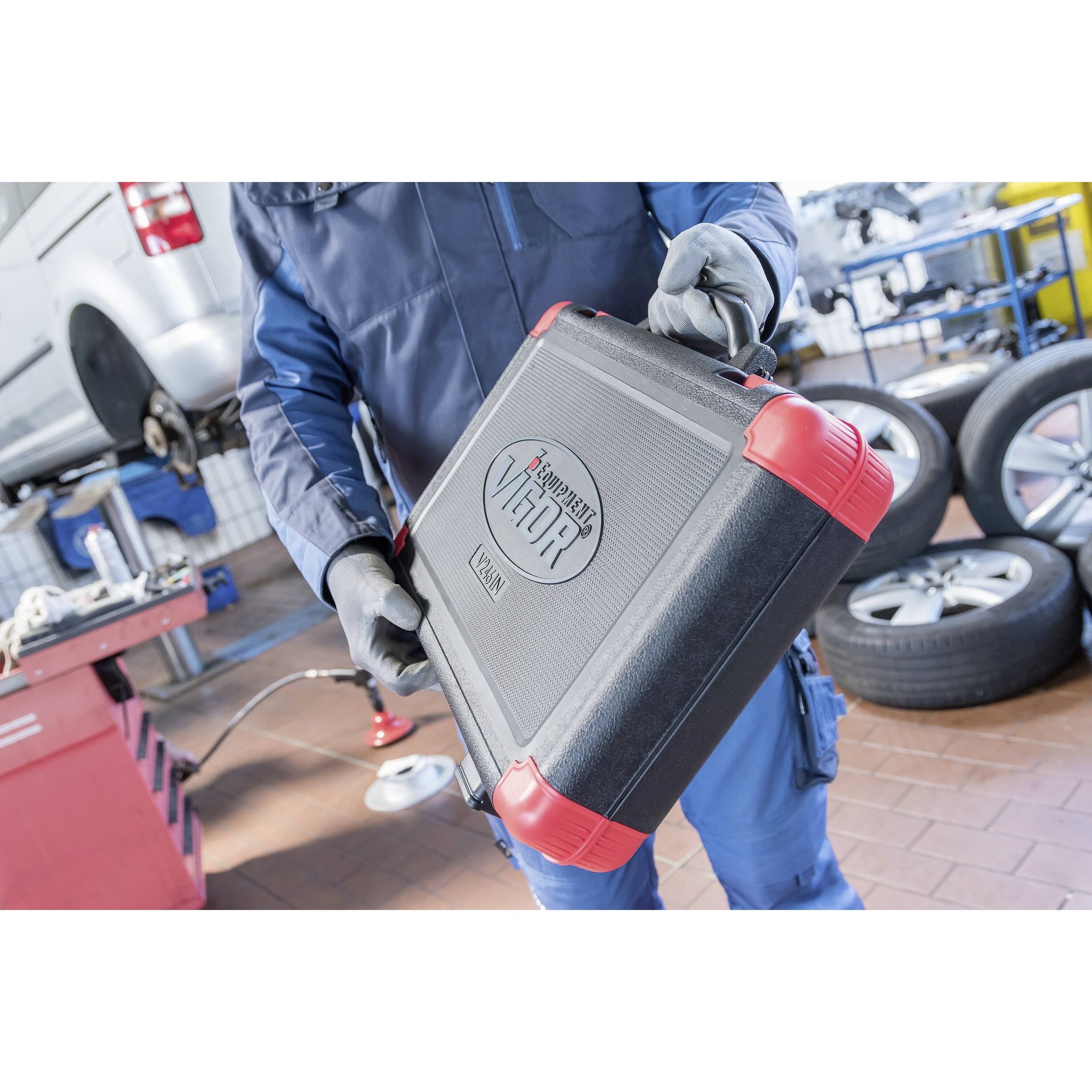 Ein Mechaniker in blauen Overalls hält einen schwarzen und roten Werkzeugkoffer mit der Aufschrift „VIGOR" in einer Autowerkstatt, mit Autos und Reifen im Hintergrund.