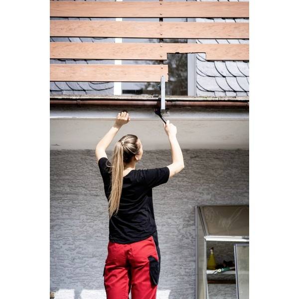 Eine Person in roten Hosen verwendet ein Werkzeug, um eine Lücke in einem Holzzaun nahe der Dachkante eines Gebäudes auszufüllen.