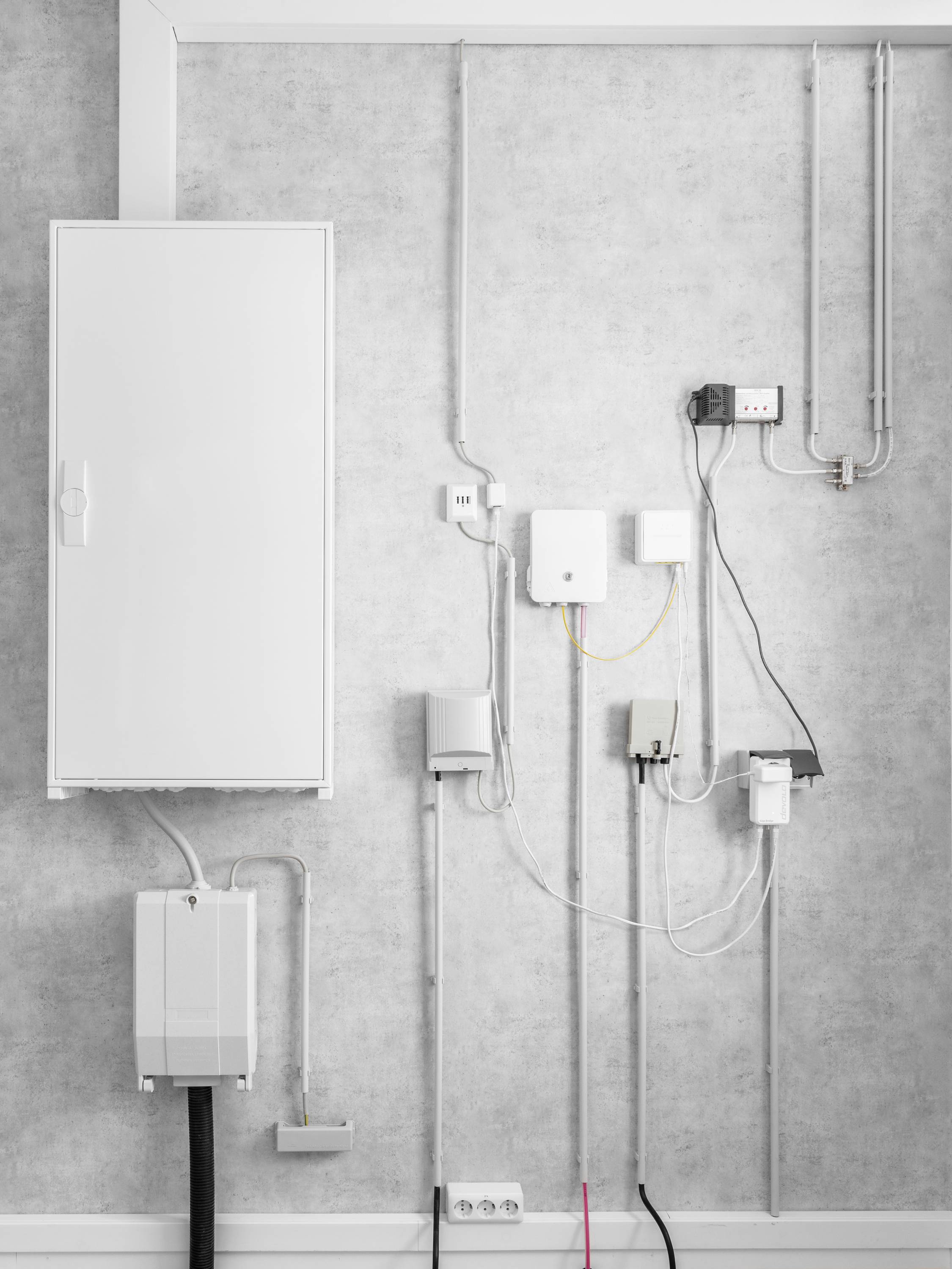 Elektrischer Schaltschrank an grauer Wand, mehrere Kabel und kleinere Kästen angeschlossen, dient zur Verwaltung elektrischer Verbindungen.