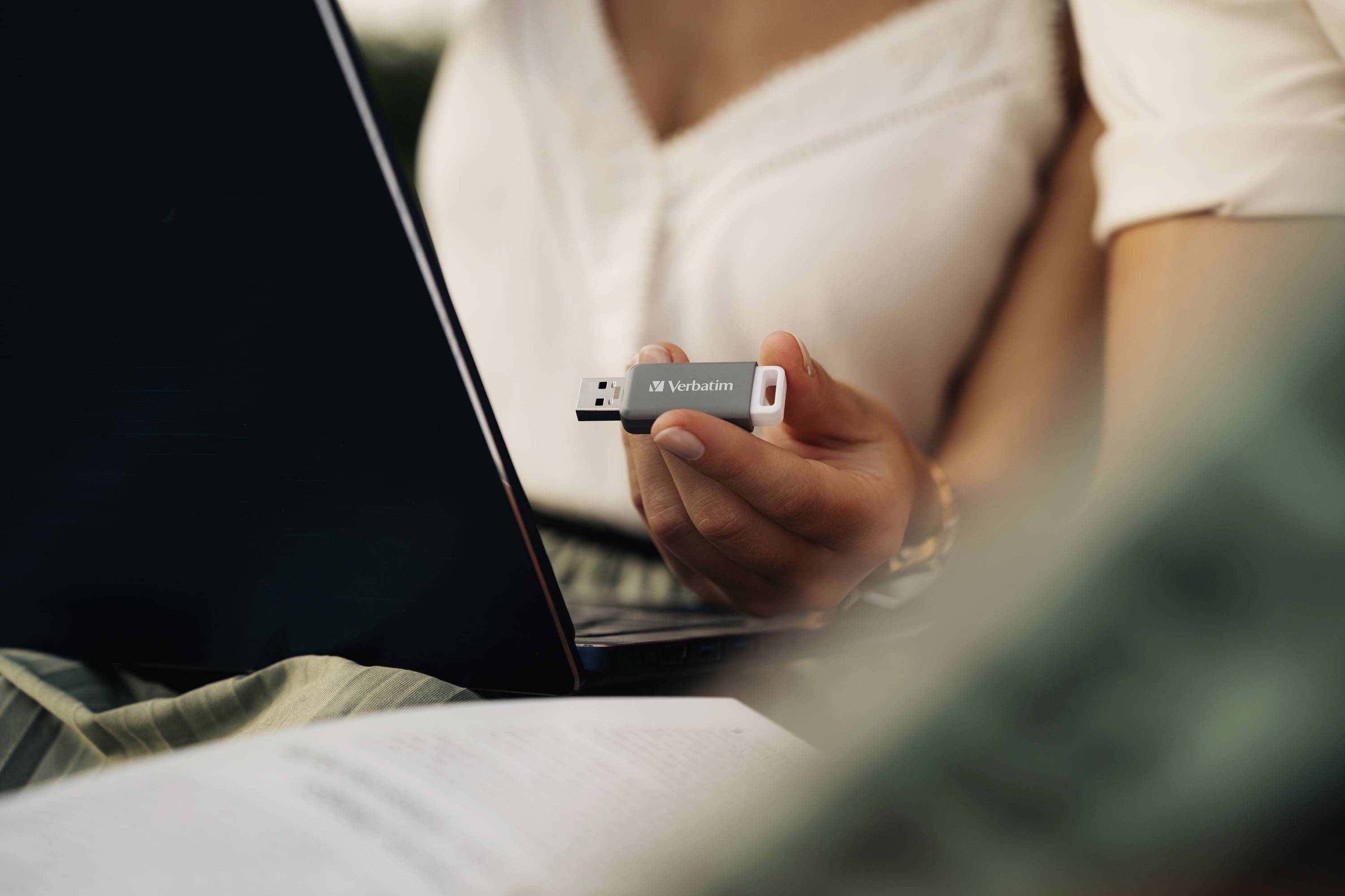 Eine Person hält einen USB-Stick und steckt ihn in einen Laptop ein. Ein offenes Buch liegt im Vordergrund.