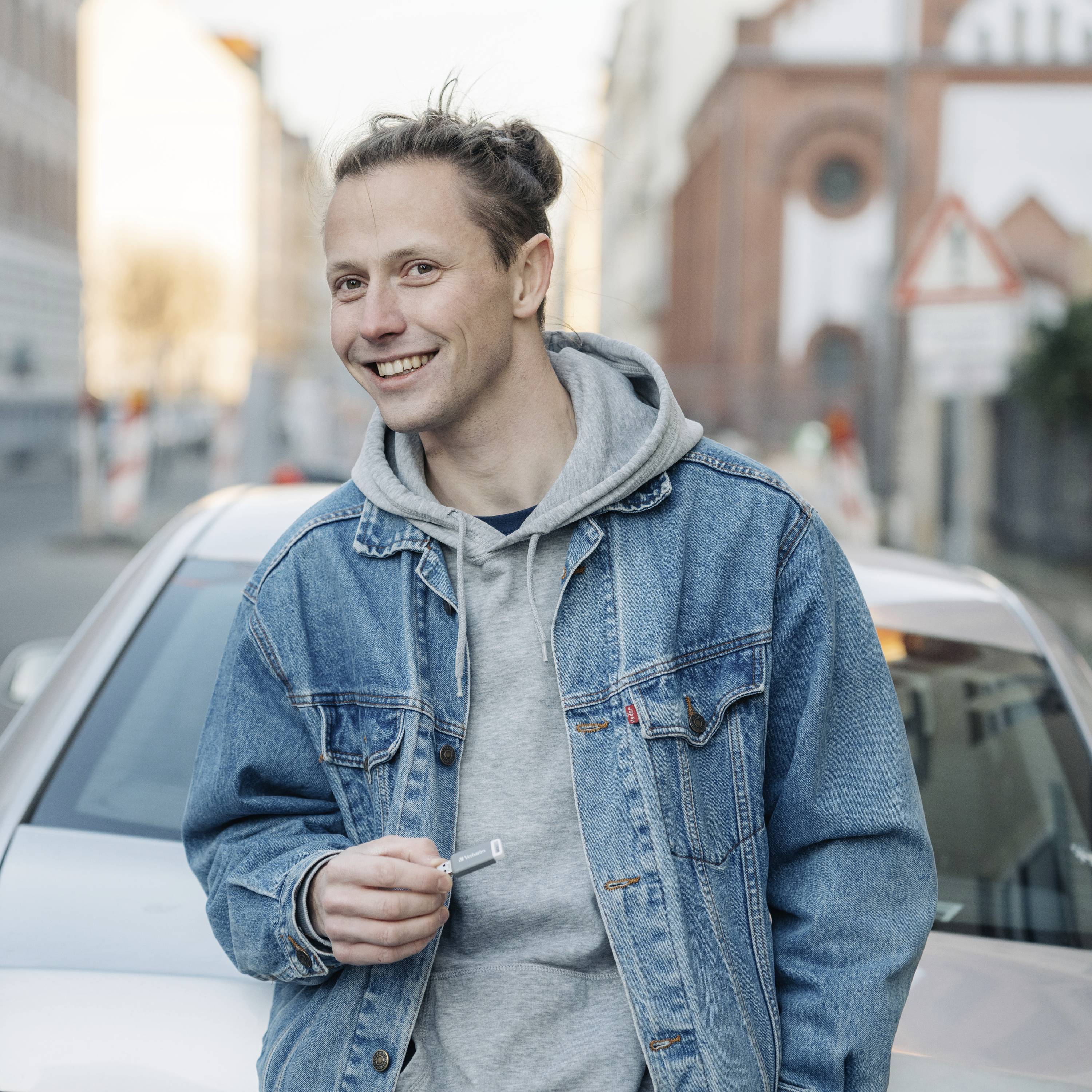 Eine Person mit Jeansjacke und grauem Hoodie steht lächelnd vor einem Auto auf einer städtischen Straße.