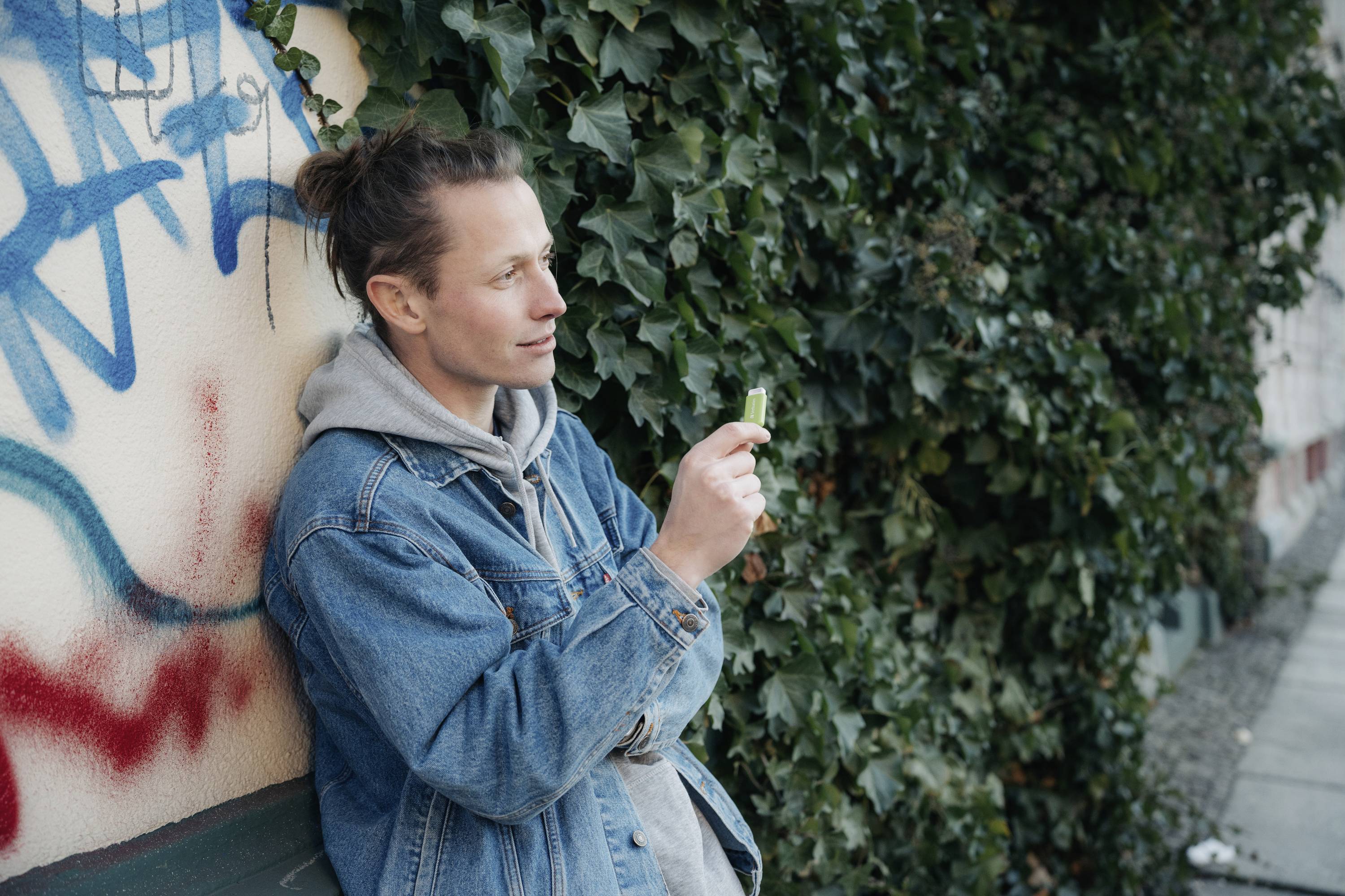 Eine Person in Jeansjacke steht an einer mit Efeu bewachsenen Wand, hält ein kleines Objekt und wirkt nachdenklich.