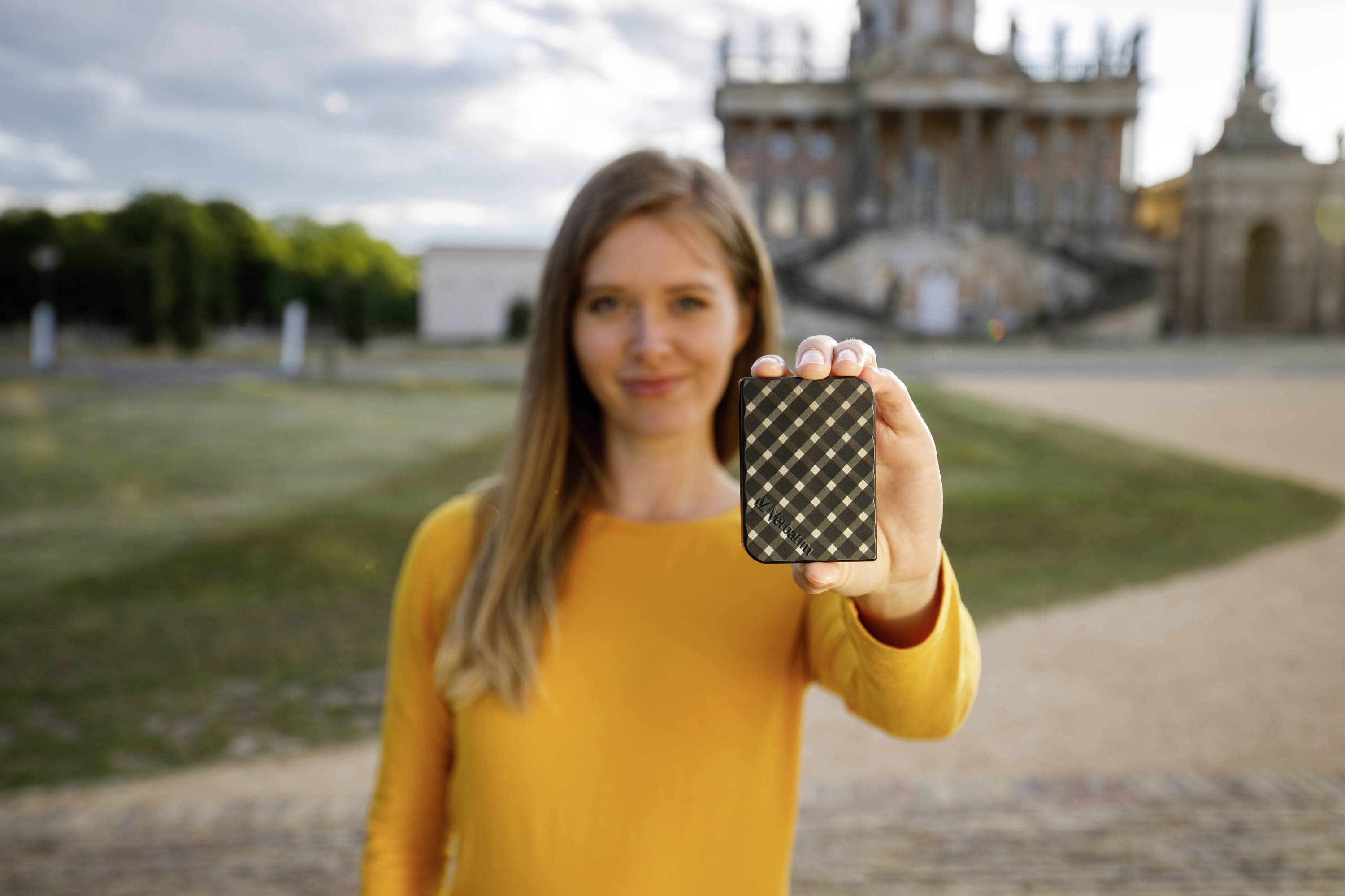 Eine Frau im gelben Pullover hält eine kleine, quadratische, schwarz-gold gemusterte Box nach vorne. Im Hintergrund ein Gebäude.