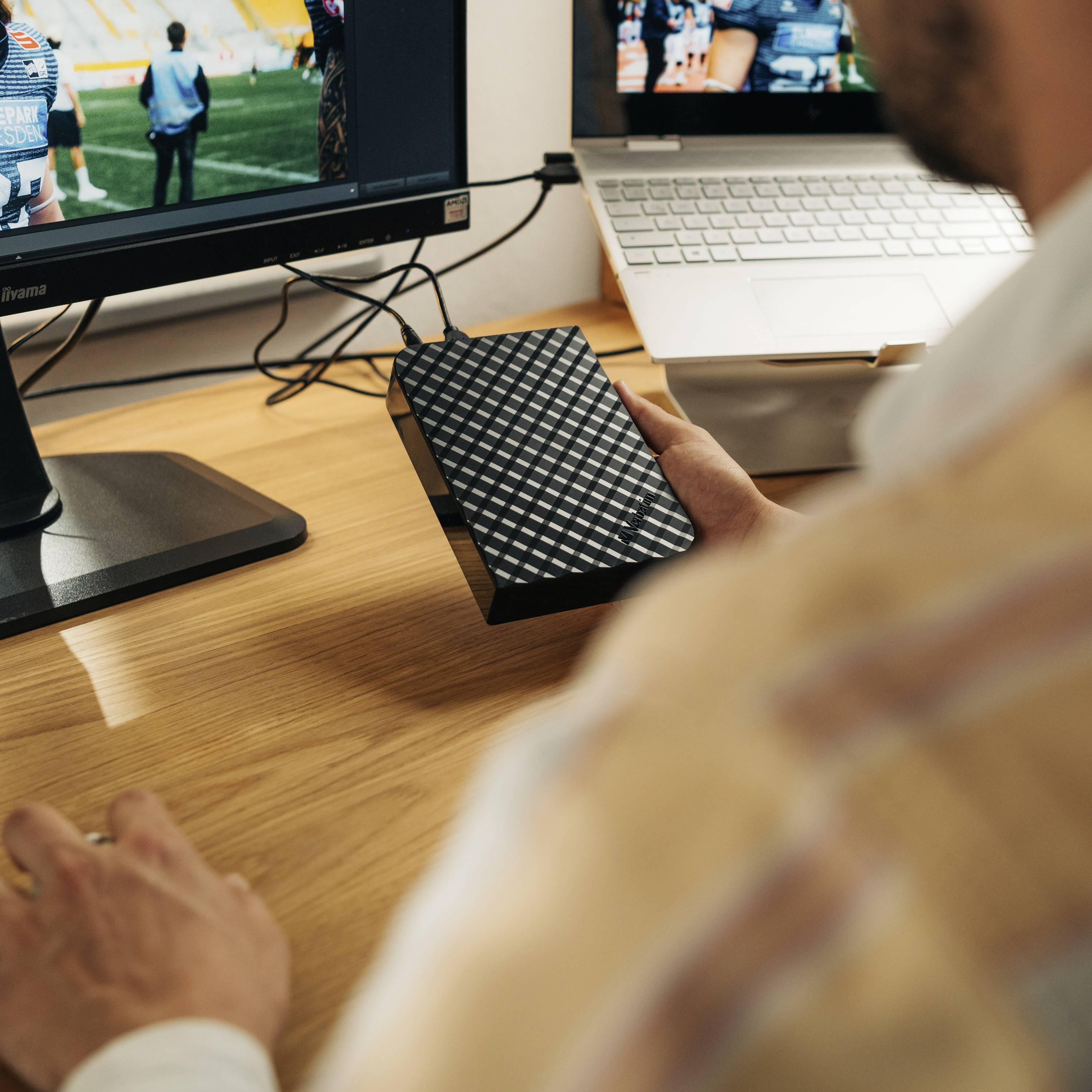 Ein Mann arbeitet an einem Computer, hält eine externe Festplatte. Auf dem Bildschirm ist ein unscharfes Sportereignis zu sehen.