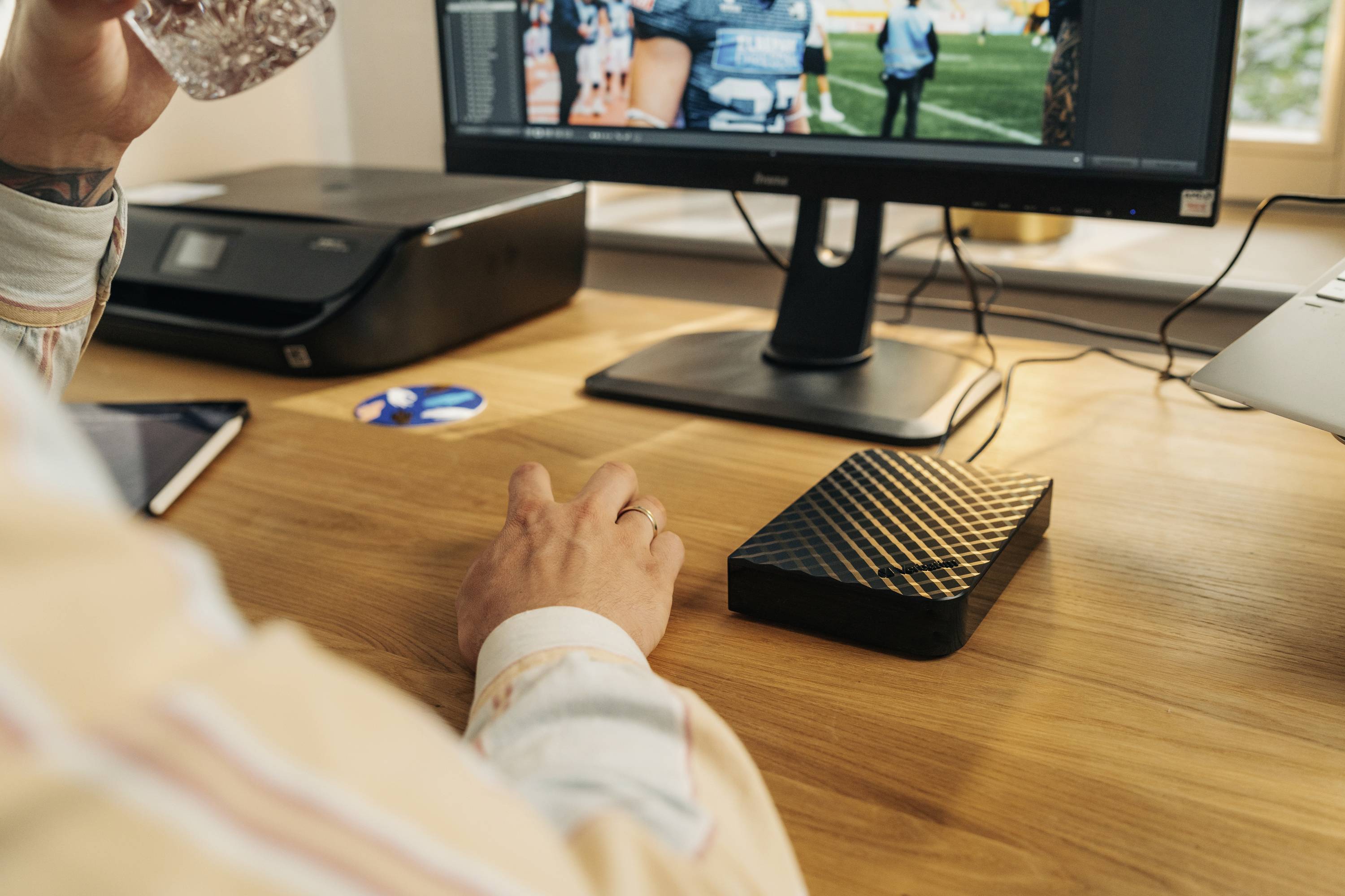 Person arbeitet am Schreibtisch mit Computerbildschirm, Getränk in der Hand. Drucker und externe Festplatte auf dem Tisch.