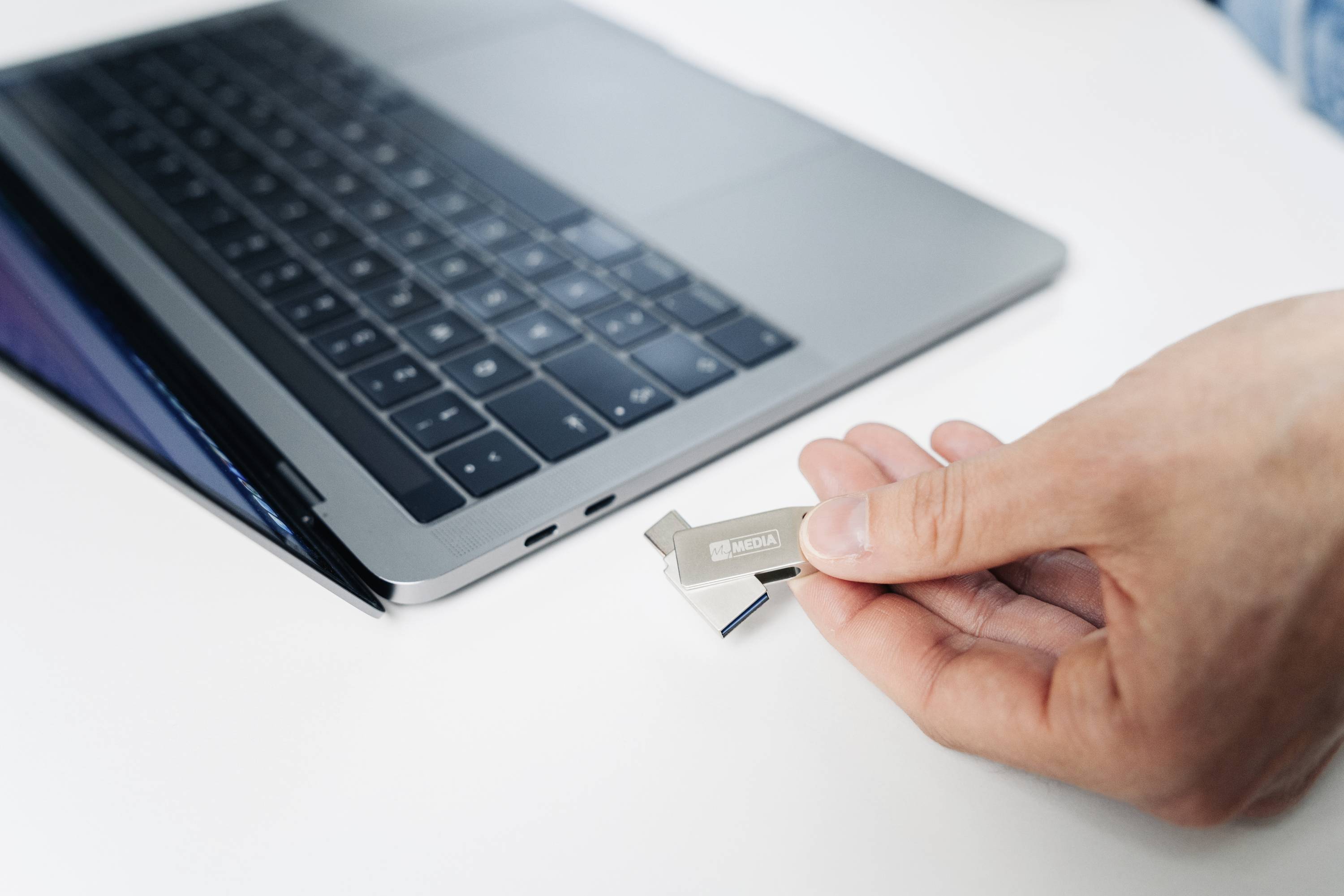 Eine Hand steckt einen USB-Stick in einen Laptop ein. Der Laptop befindet sich auf einem weißen Tisch und zeigt eine teilweise geöffnete Ansicht.