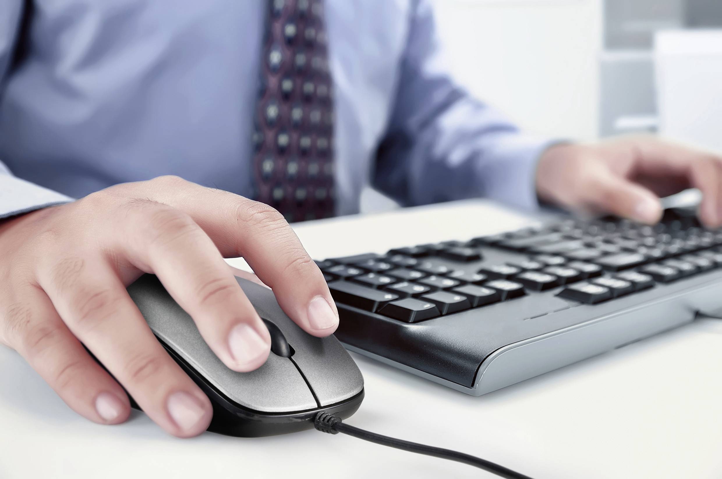 Eine Person arbeitet am Computer, sieht auf den Bildschirm, während sie eine Maus in der rechten Hand hält und die linke auf der Tastatur hat.