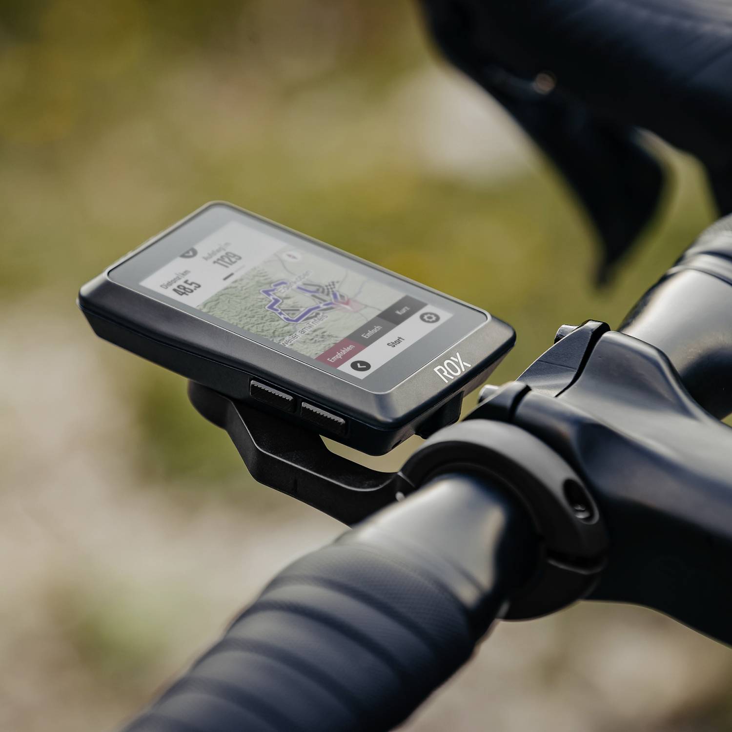 Ein Fahrradcomputer auf einem Lenker zeigt eine Karte und Daten wie Geschwindigkeit, Trittfrequenz und Uhrzeit an. Im Hintergrund unscharf.