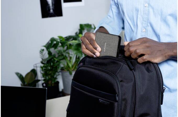 Person packt eine graue Seagate-Festplatte in einen schwarzen Rucksack. Im Hintergrund sind ein Schreibtisch, Pflanzen und Bilder an der Wand zu sehen.