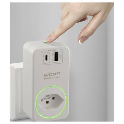 Eine Hand drückt einen Knopf auf einem Voltcraft USB-Ladegerät, das in eine Steckdose gesteckt ist, mit zwei USB-Anschlüssen und einer Steckdose.